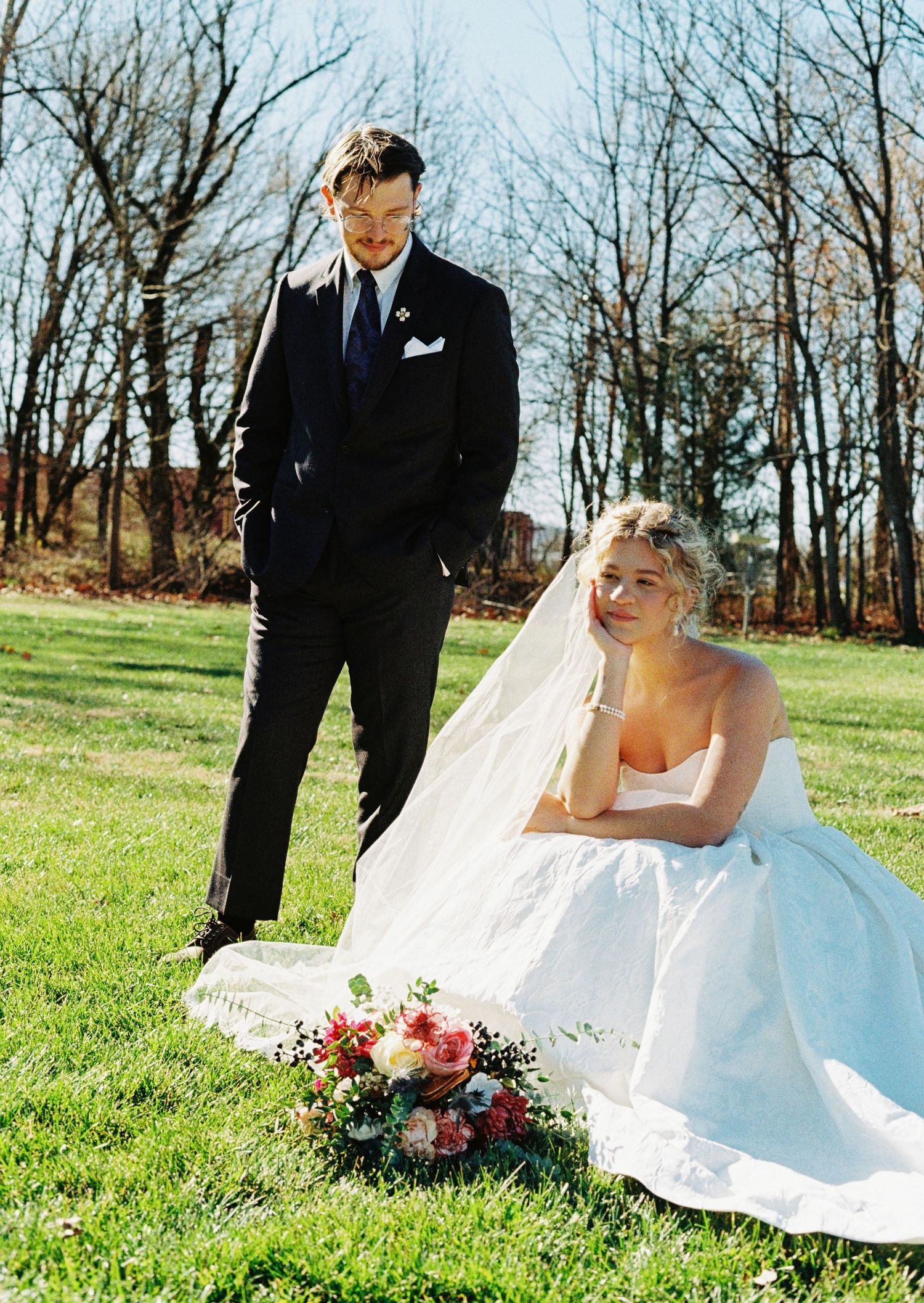Bride in white gown sits on grass holding bouquet while groom in dark suit stands beside her outdoors.