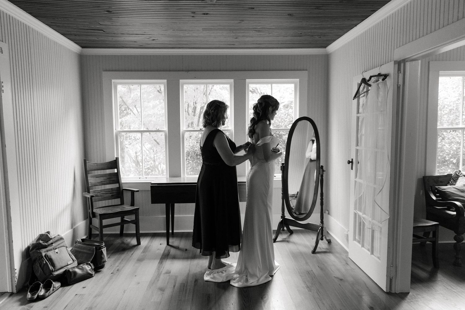 Black and white photo of a bride being helped into her wedding dress by a bridesmaid in a sunlit room with a mirror.