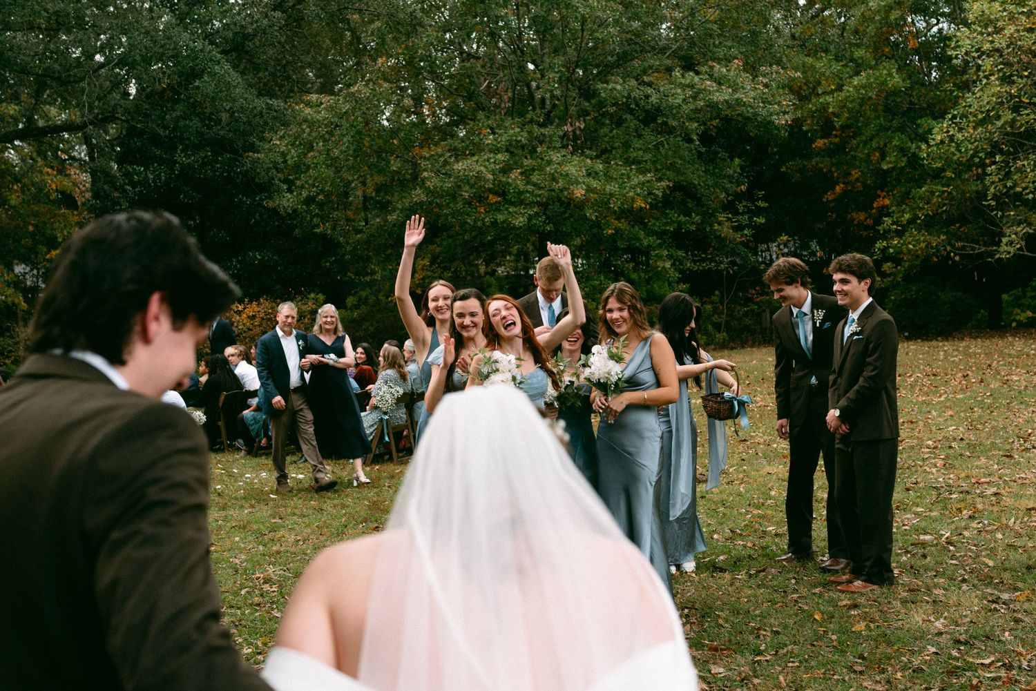 Bride and bridesmaids in blue dresses celebrate joyfully outdoors at a fall wedding ceremony surrounded by lush green trees.