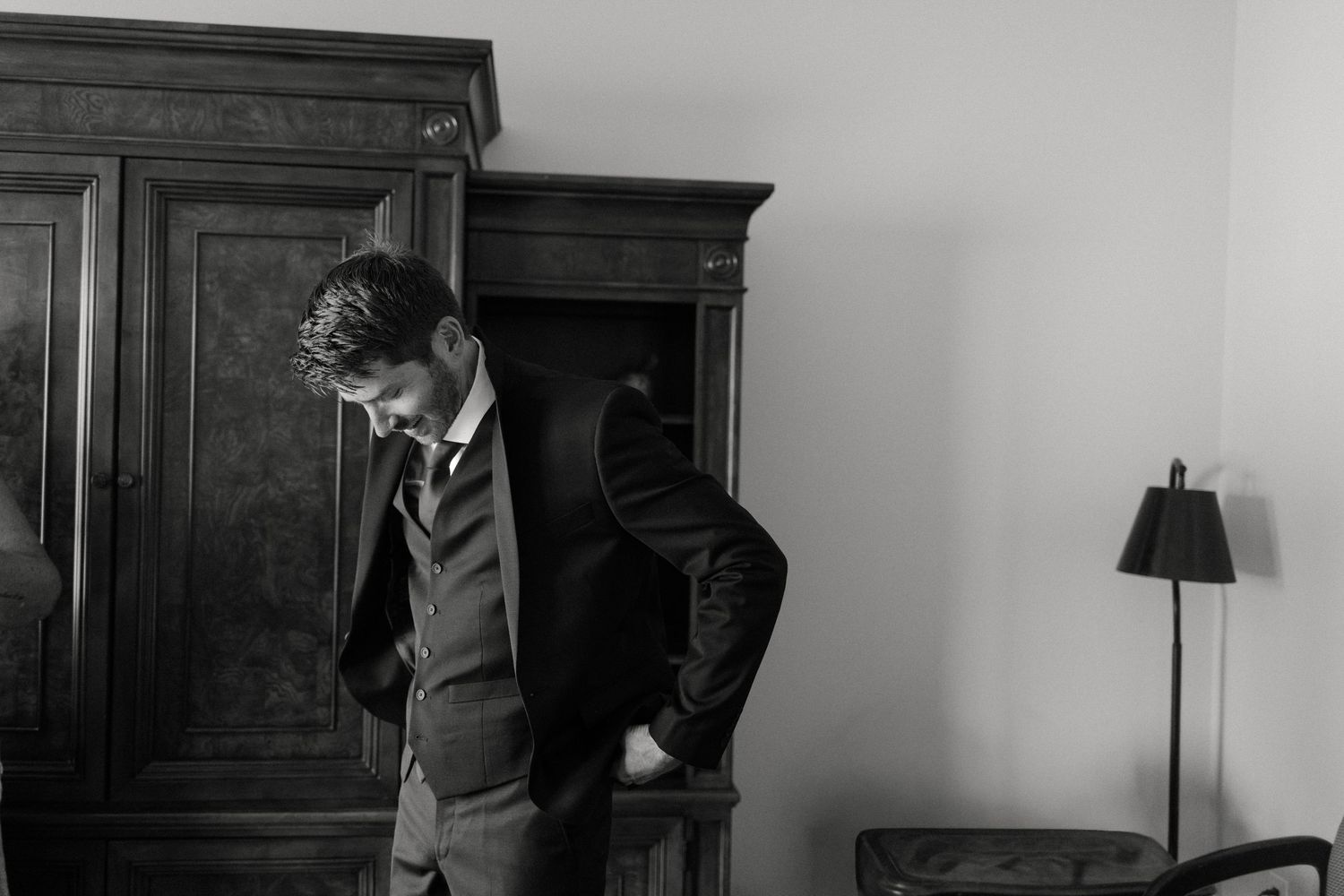 Black and white photo of a groom in a vest and tie adjusting his suit jacket beside a wooden armoire.