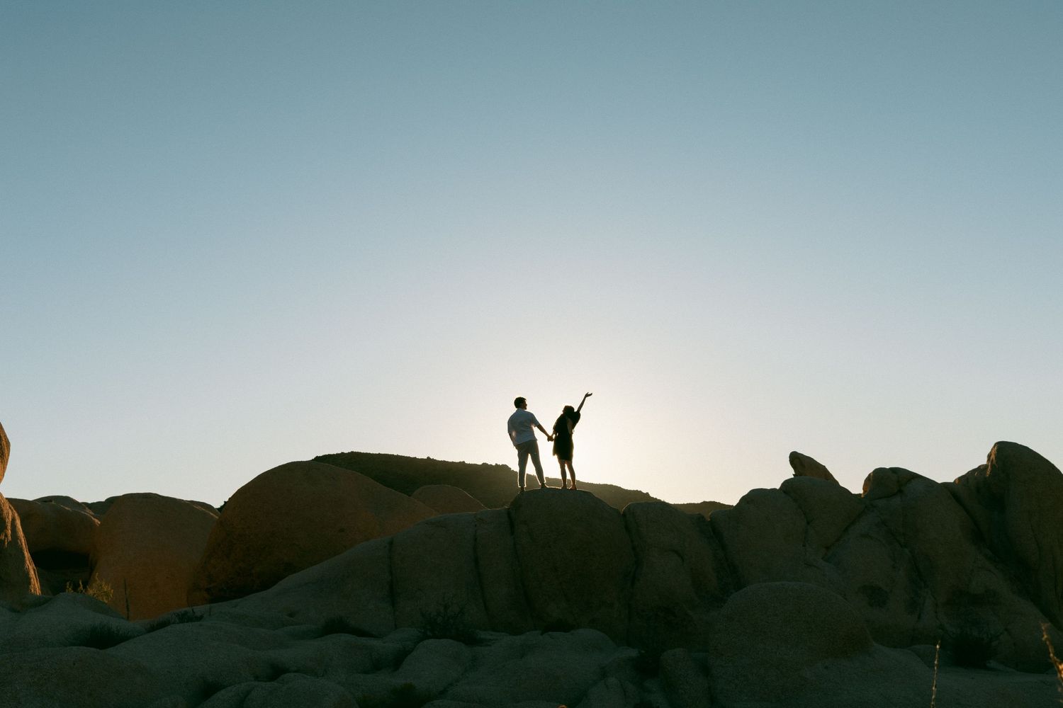 Two silhouetted figures stand atop rocky boulders against a golden sunset sky in a desert landscape.
