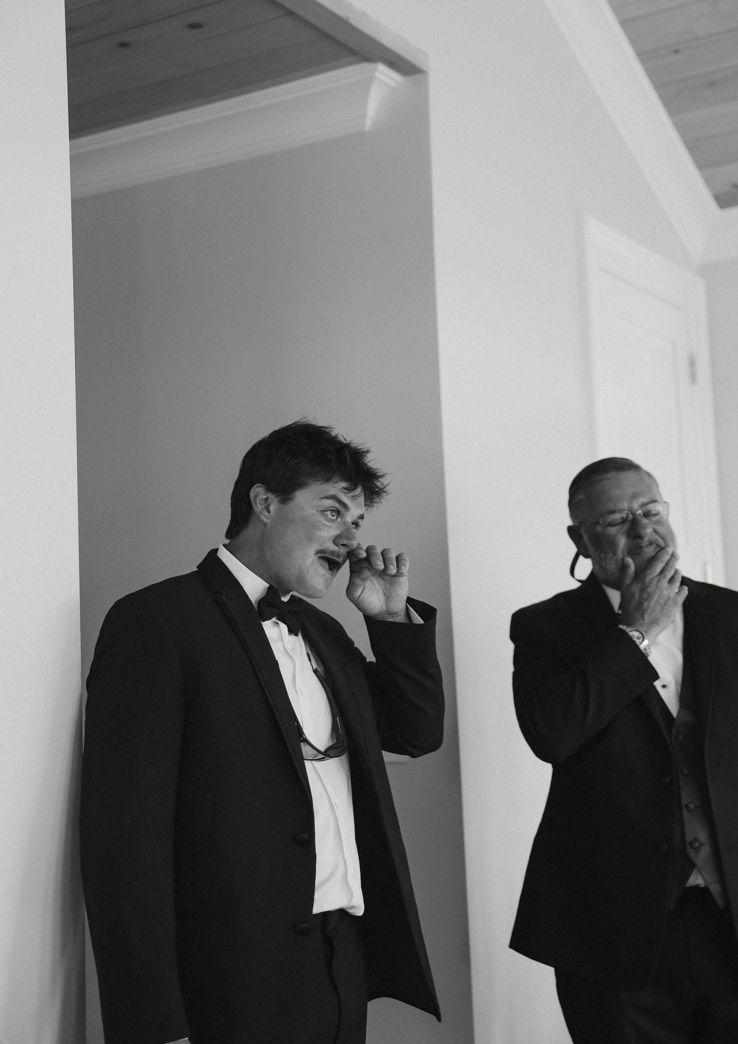 Two men in formal suits emotional at a wedding, one wiping tears, black and white photo.