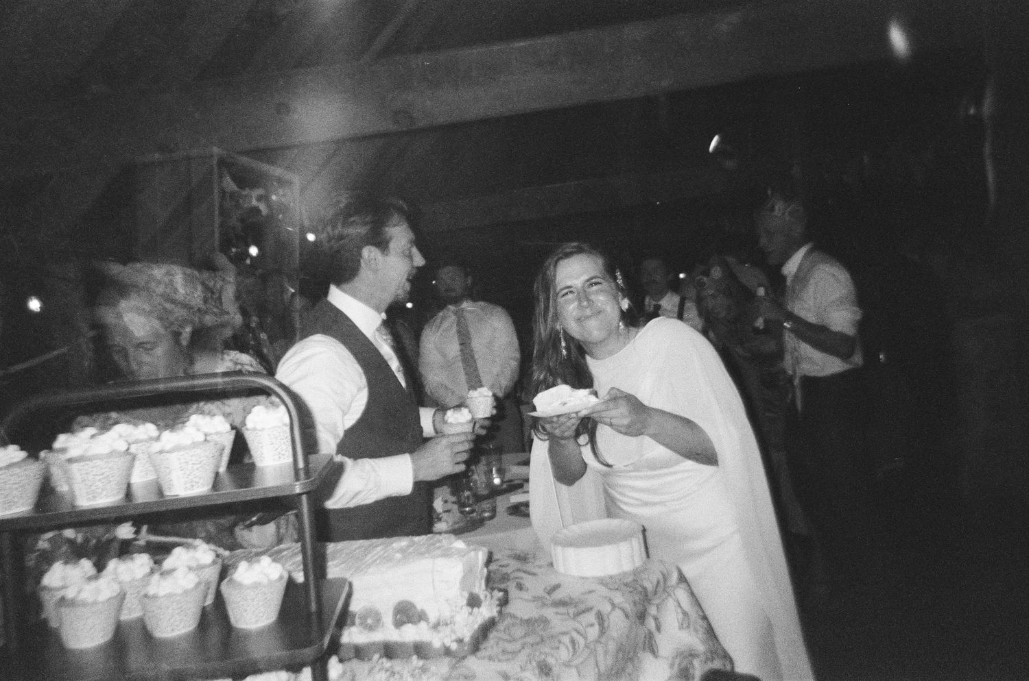 Black and white photo of a bride and groom smiling at a wedding reception dessert table with cupcakes.