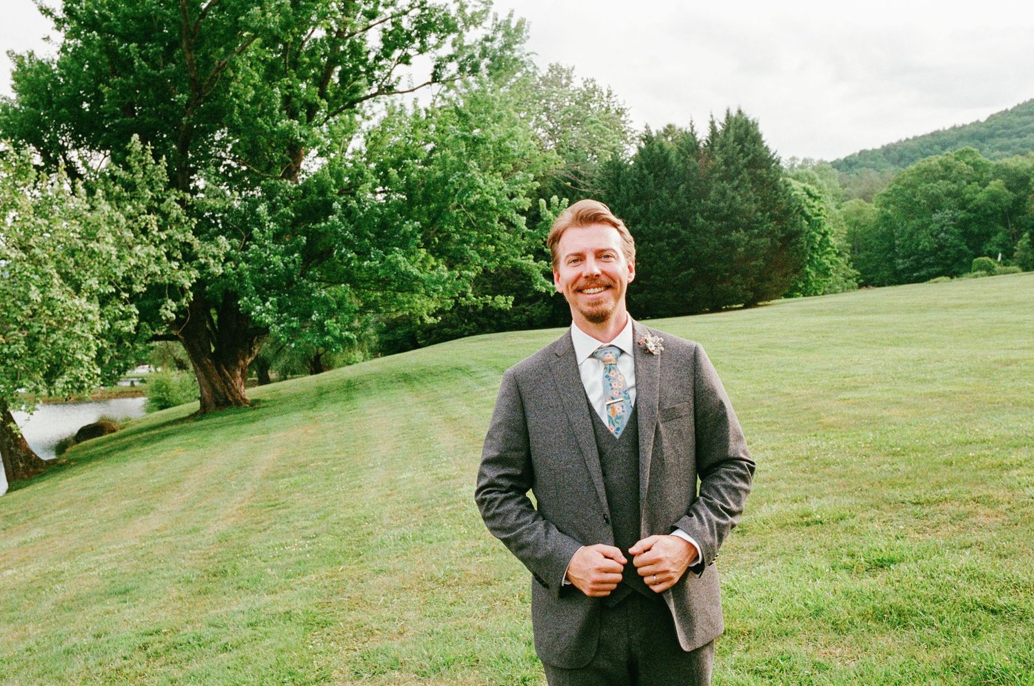 Smiling groom in gray suit adjusting jacket buttons outdoors on green lawn with trees and hills in background.