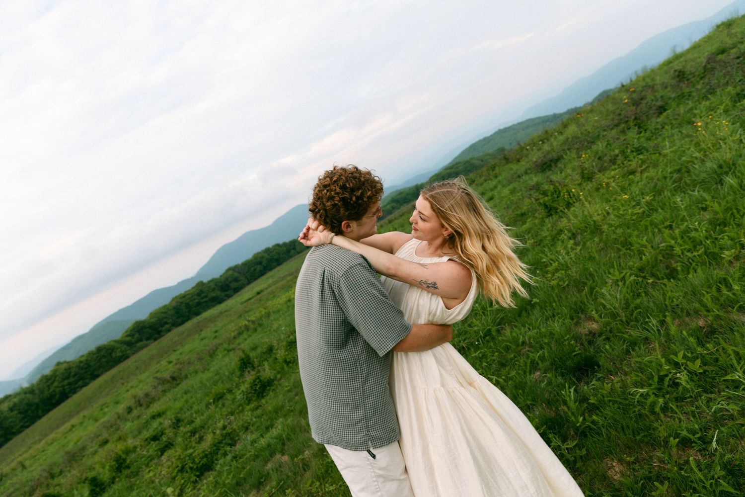Couple dancing and embracing on green hilltop meadow with scenic mountain range in the background.