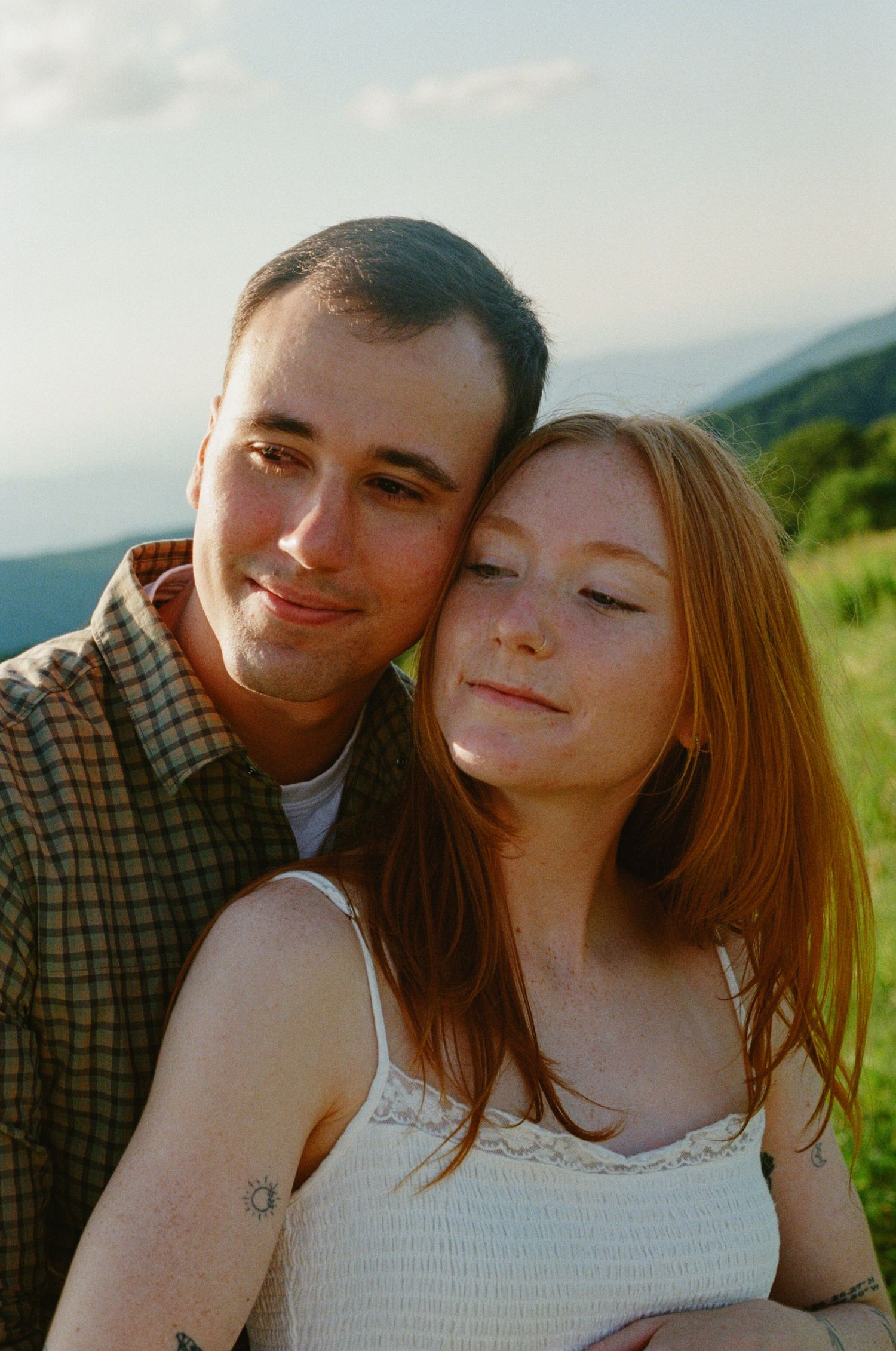 A young couple poses outdoors, the woman with red hair leaning back against a man, both smiling with mountains behind.