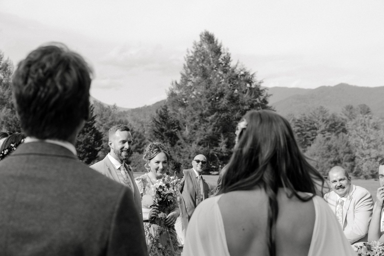 Black and white outdoor wedding ceremony with guests smiling as bride approaches groom, mountains and trees in background.
