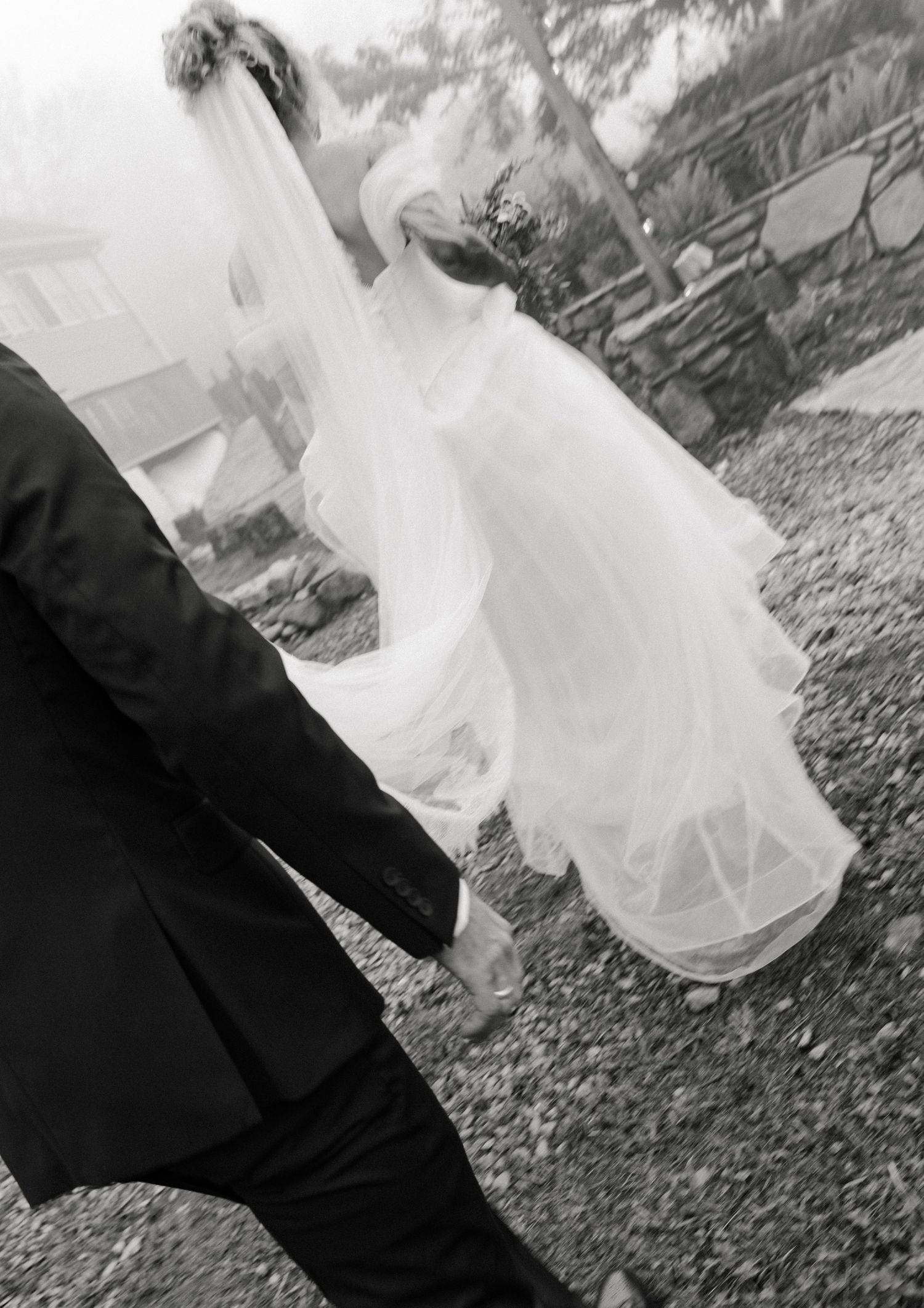 Black and white photo of bride in flowing veil and gown walking hand in hand with groom in dark suit.