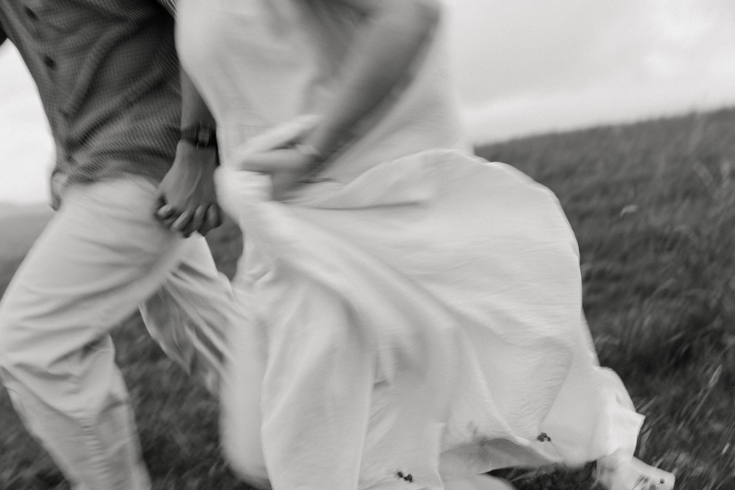 Black and white photo of a couple holding hands, walking through a grassy field, dress flowing in the wind.