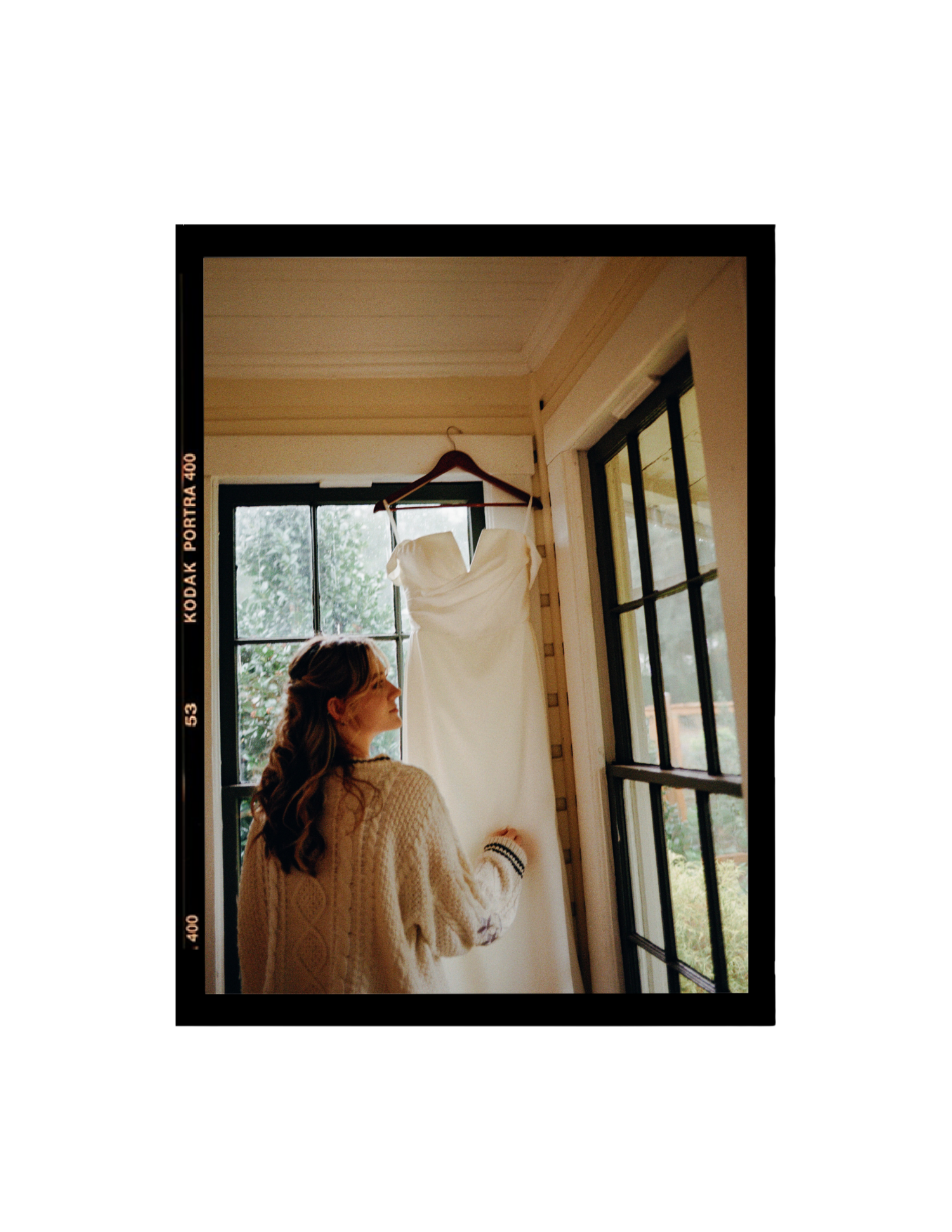 Bride admiring her white wedding dress hanging by a window, captured on Kodak Portra 400 film.