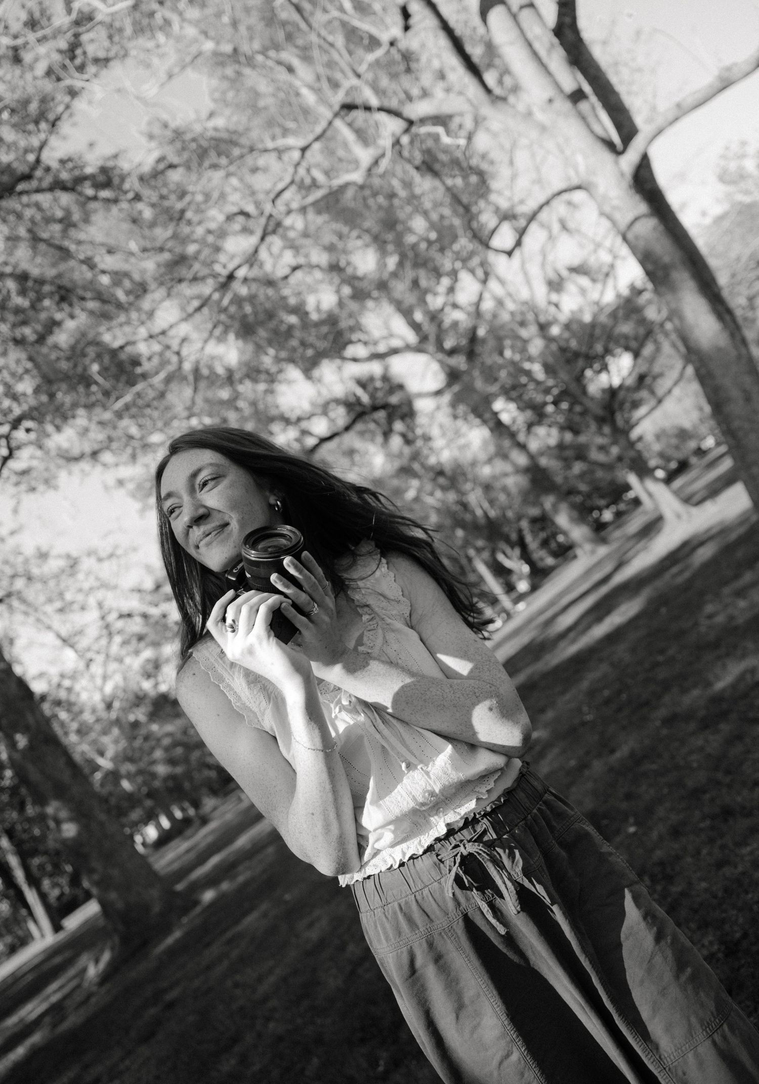 Black and white photo of a smiling young woman holding headphones in a sunlit park with trees in the background.