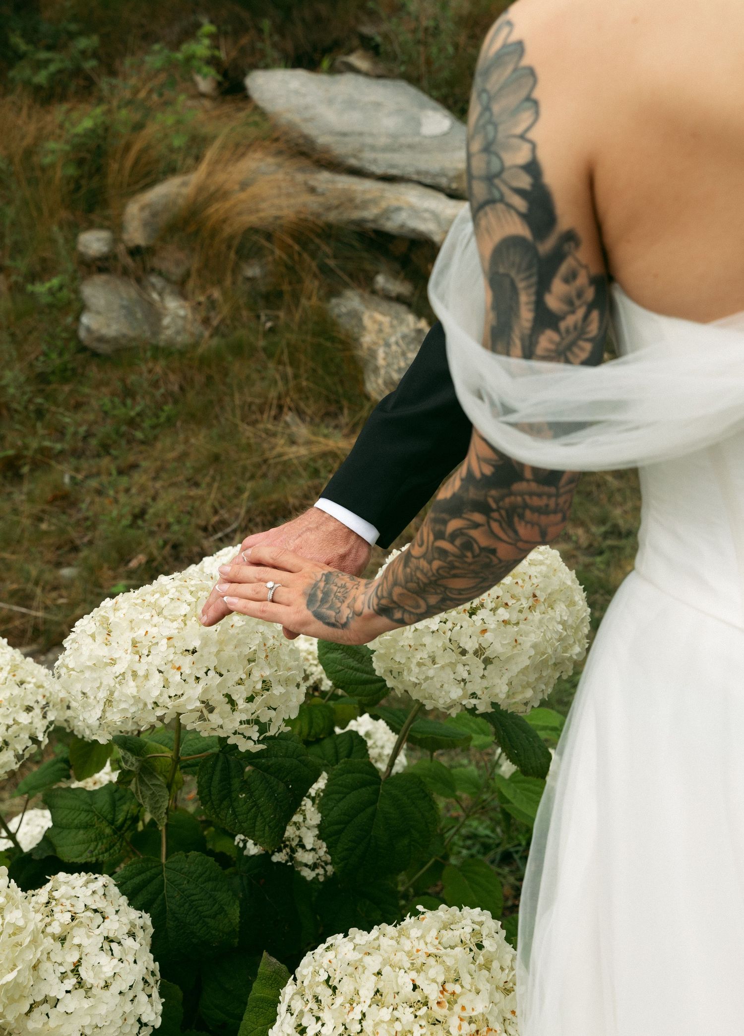 Tattooed bride and groom holding hands over white hydrangea flowers in an outdoor wedding setting.