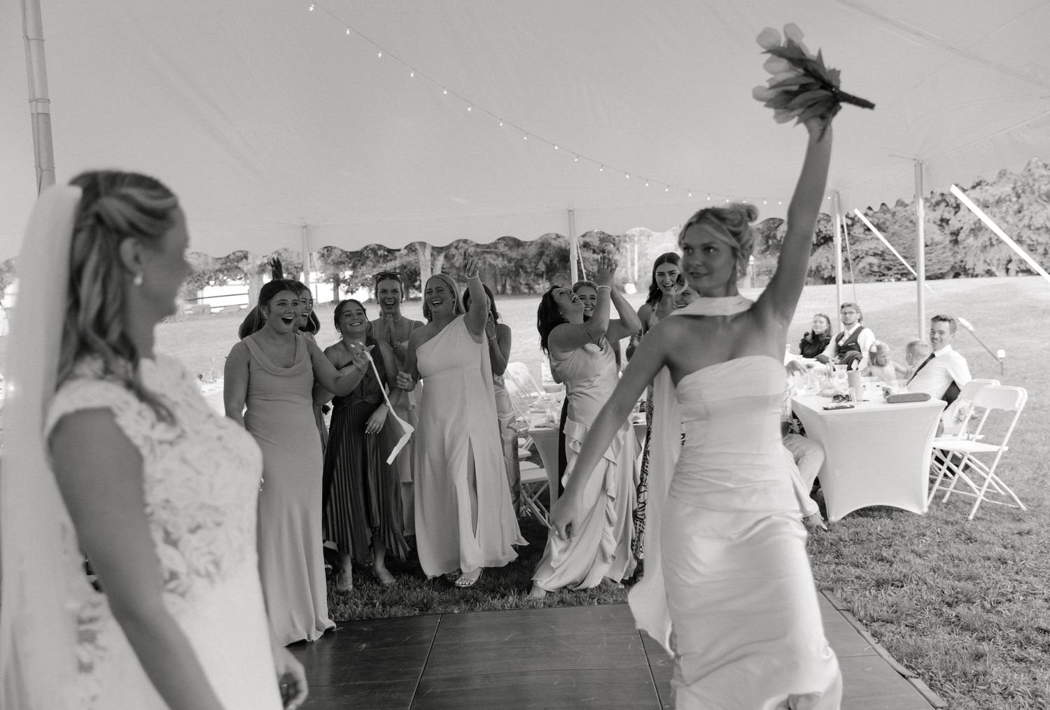 Bride tossing bouquet to excited bridesmaids at outdoor wedding reception under tent in black and white photo.