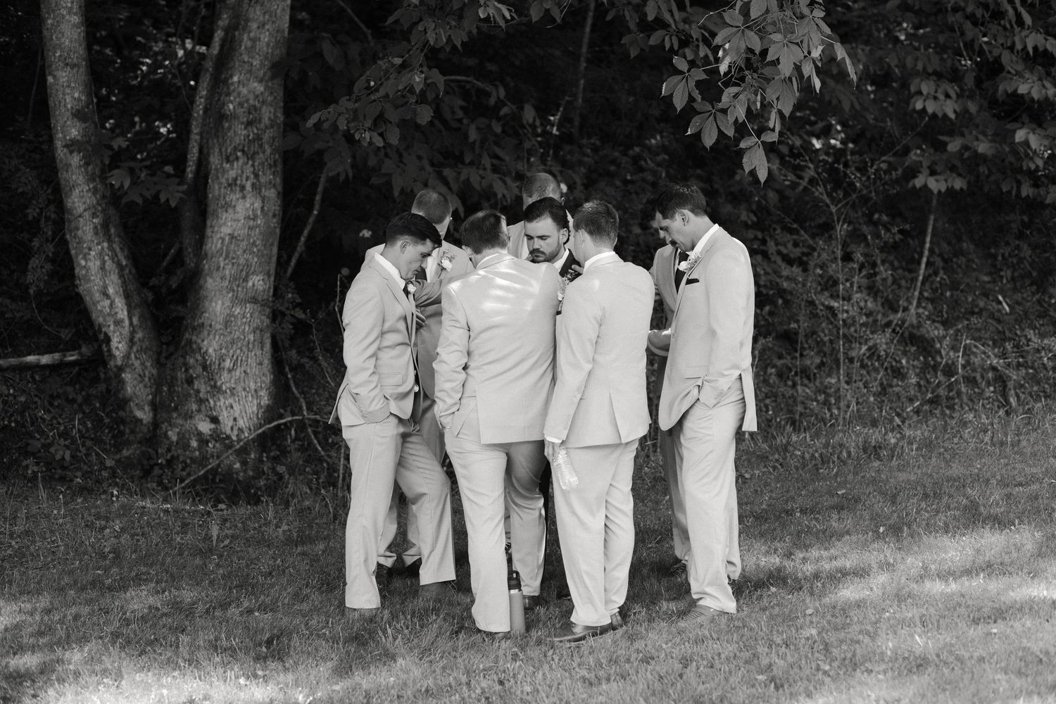 Black and white photo of groomsmen in suits huddled together outdoors near trees on a wedding day.