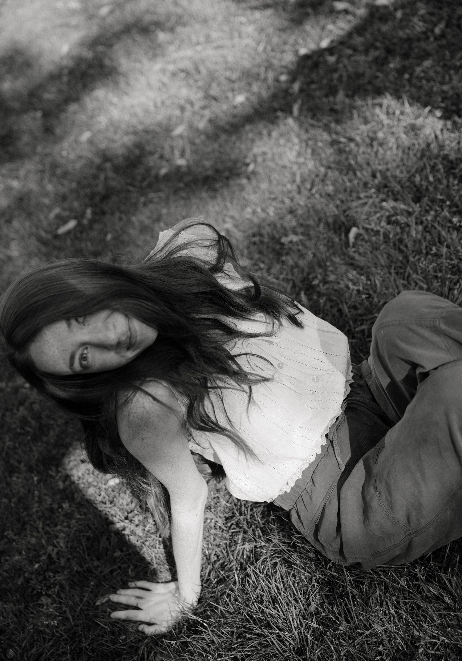 Black and white photo of a young woman with long hair lying on grass, wearing a white top and dark pants.