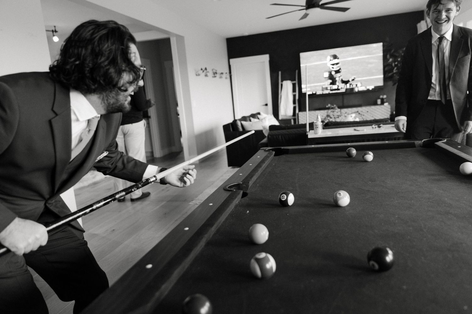 Black and white photo of a man in a suit playing pool at a billiard table with others watching nearby.