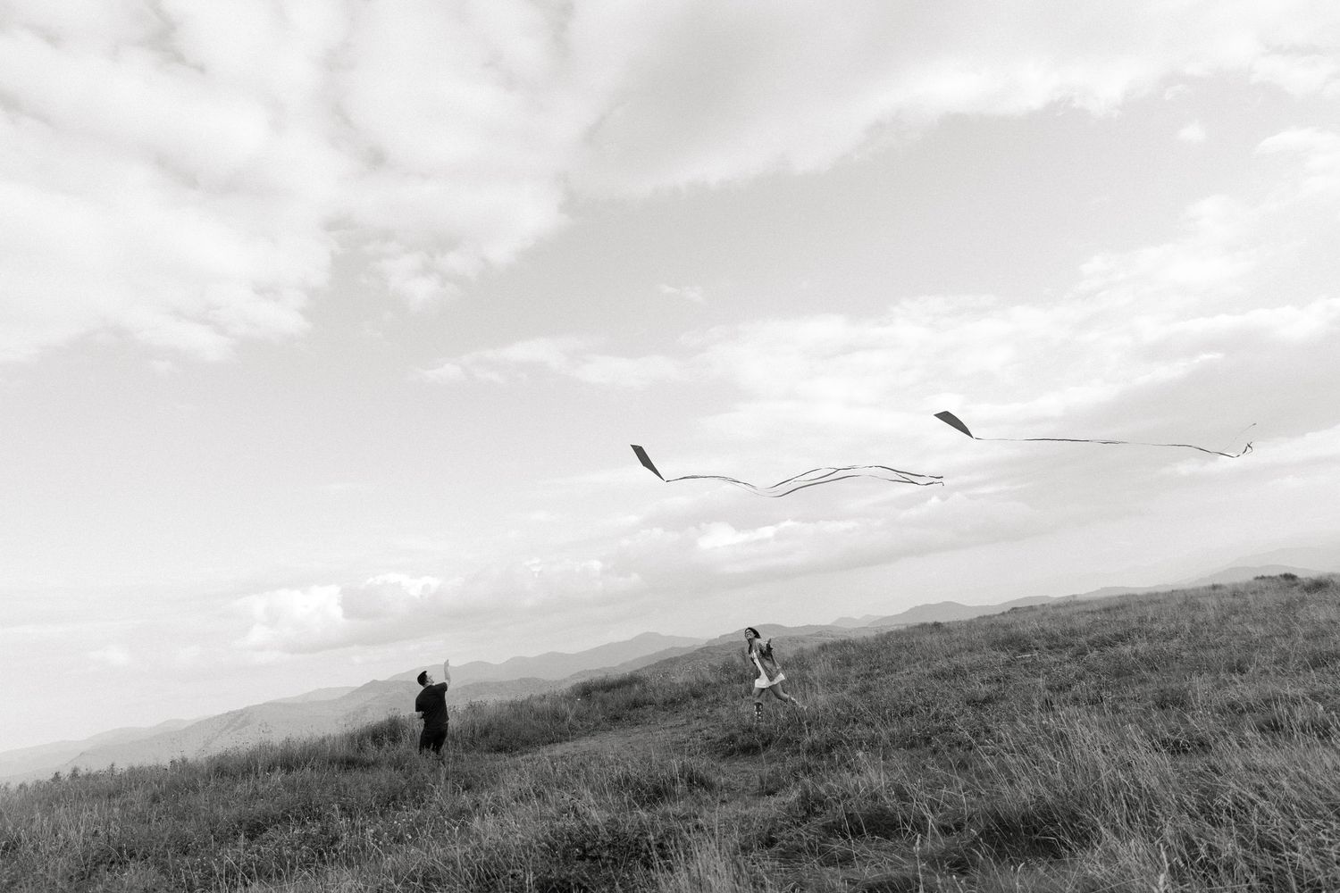 Two people flying kites on a grassy hillside under a cloudy sky in a black and white landscape photo.