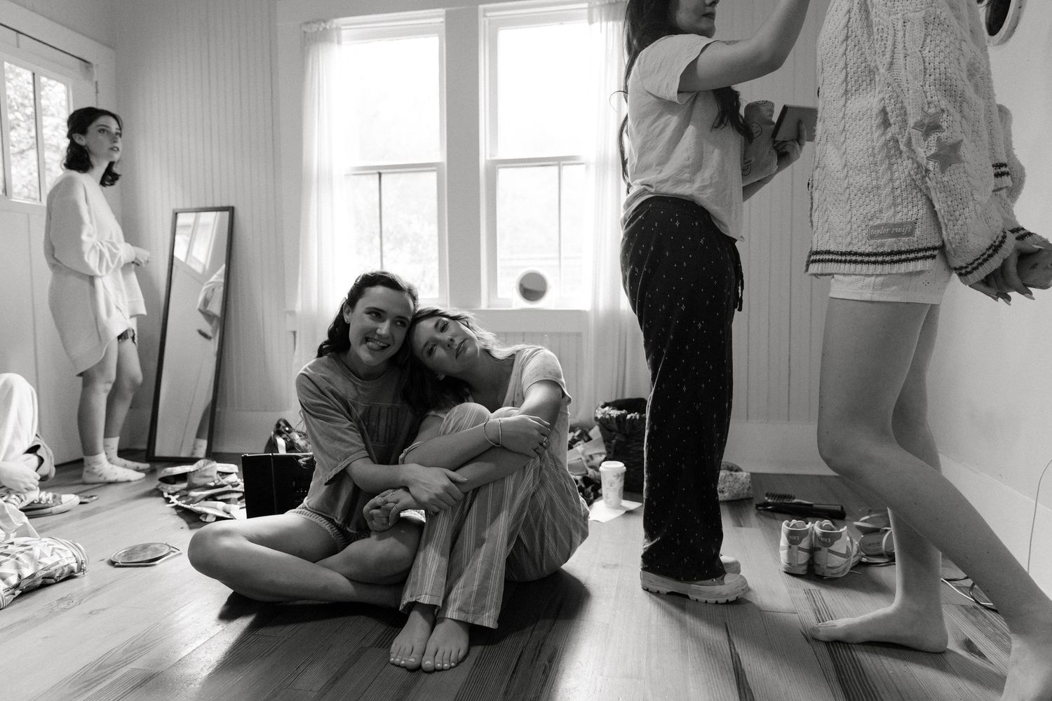 Black and white photo of people relaxing in a bright room with hardwood floors, two sitting on the floor embracing.