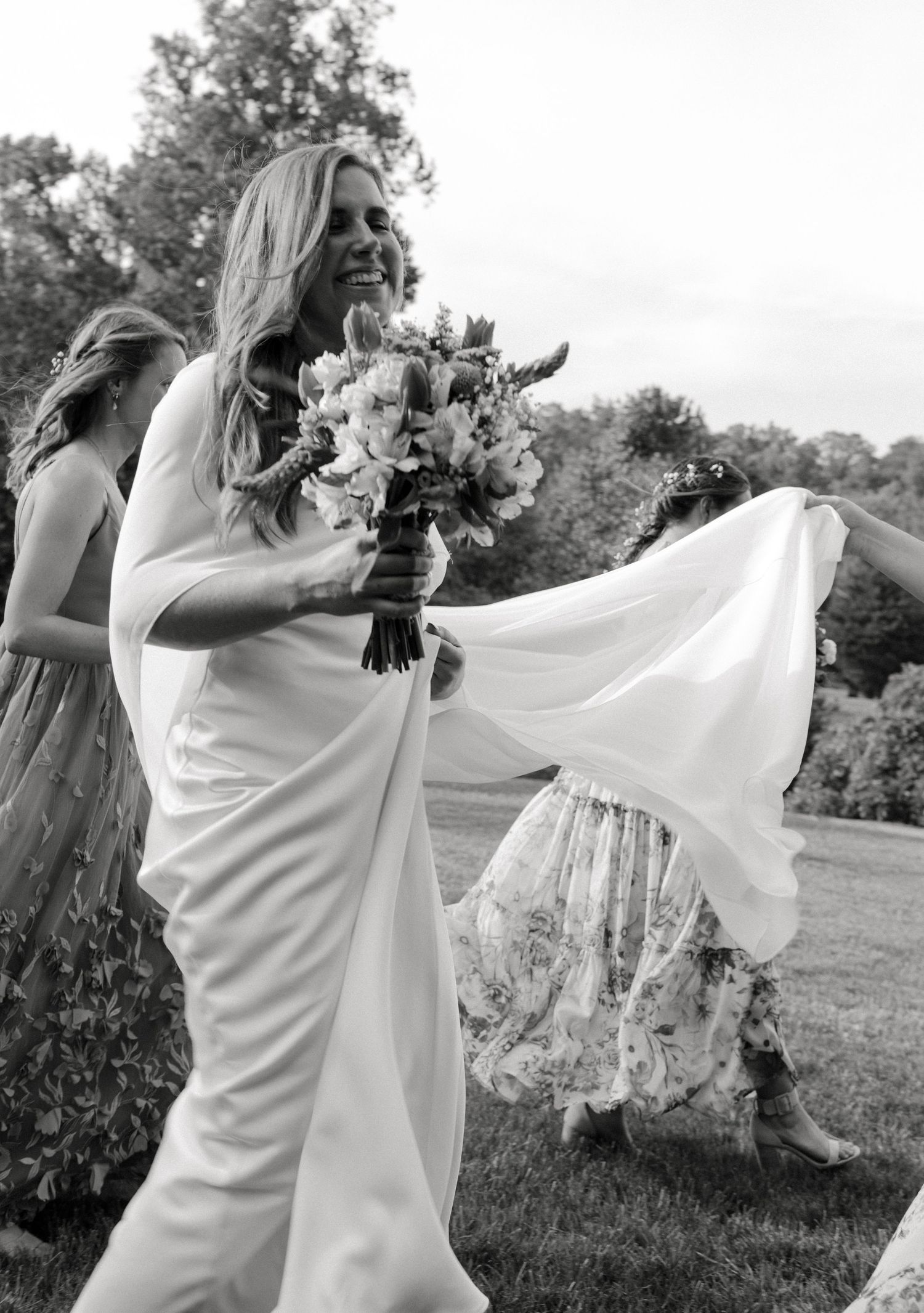 Black and white photo of a smiling blonde bride in white gown carrying her bouquet outdoors.