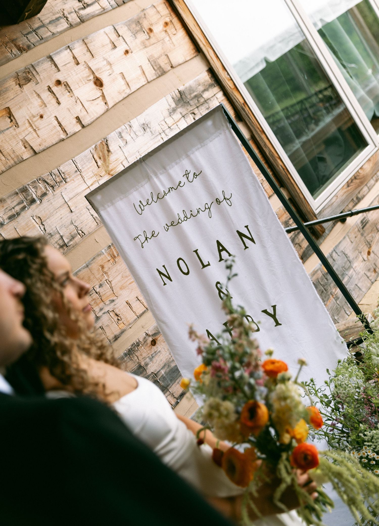 Couple stands before a white wedding banner reading Welcome to the Wedding of Nolan and Abby with colorful floral arrangements.