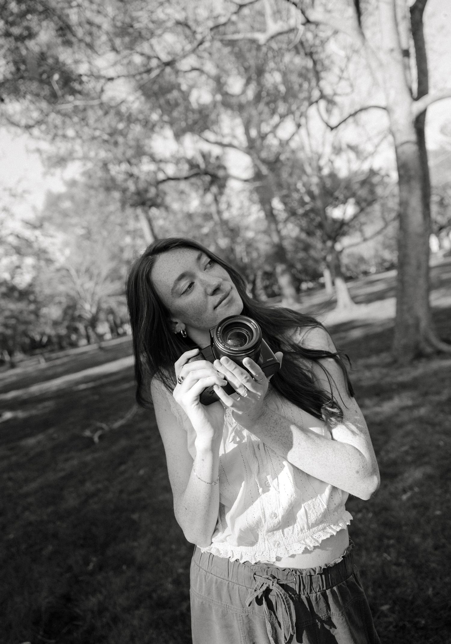 Black and white photo of a young woman holding a vintage camera in a park, eyes closed, wearing a white top.