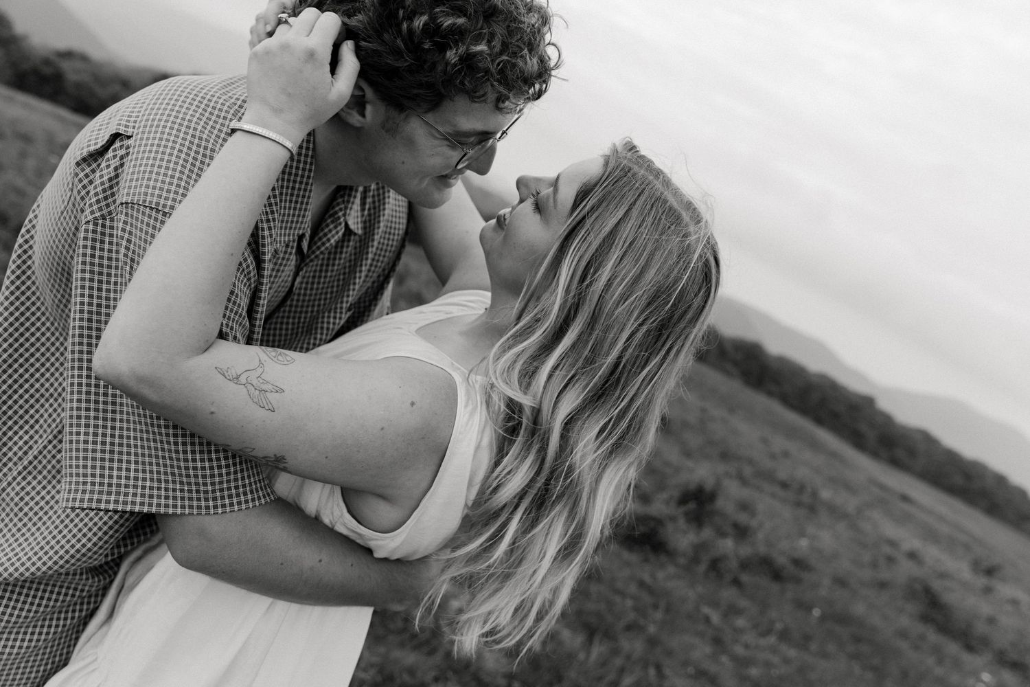 Black and white photo of a couple sharing a romantic kiss in an open field with mountains in the background.