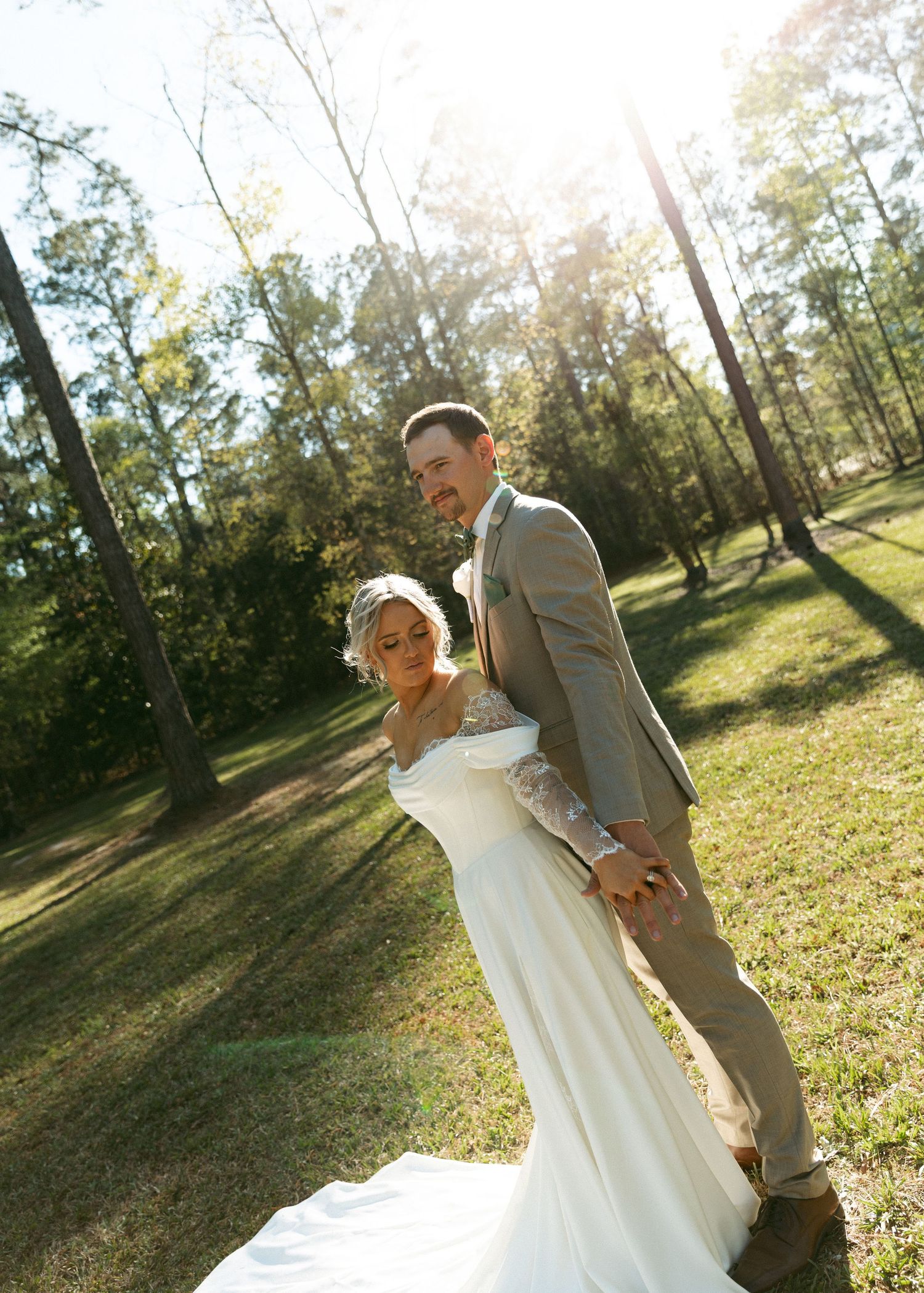 Bride in white gown and groom in gray suit hold hands in sunlit forest clearing during outdoor wedding portrait.