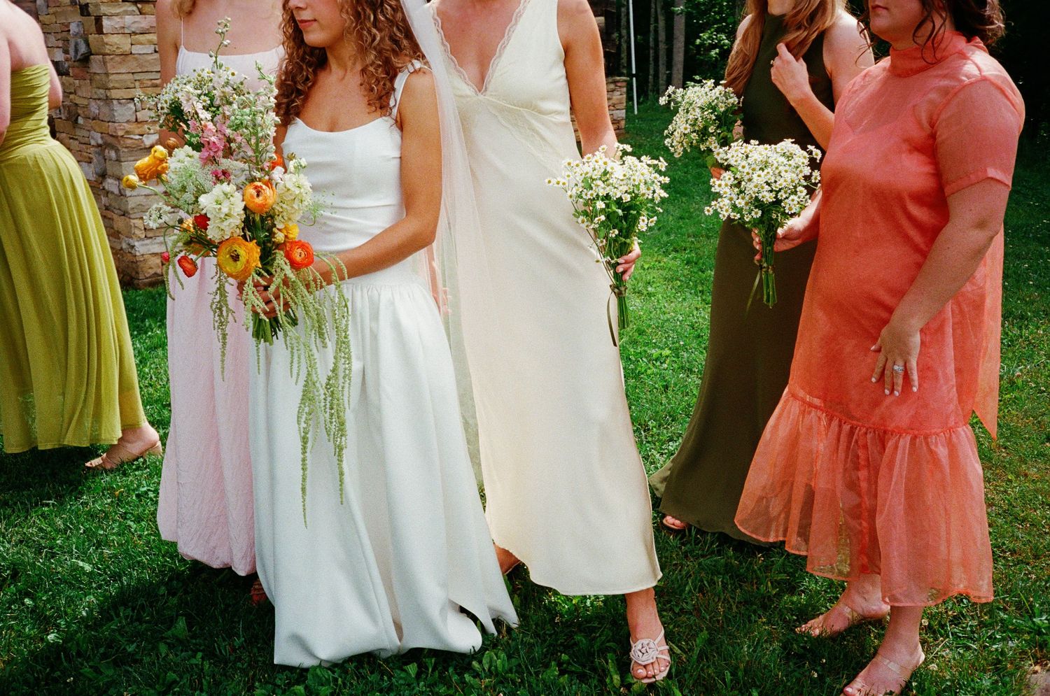 Bride in white gown with bridesmaids in colorful dresses holding bouquets of wildflowers outdoors.