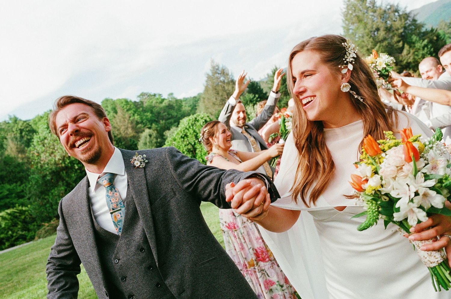 Joyful newlyweds hold hands outdoors, bride holding white floral bouquet, guests celebrating behind them.