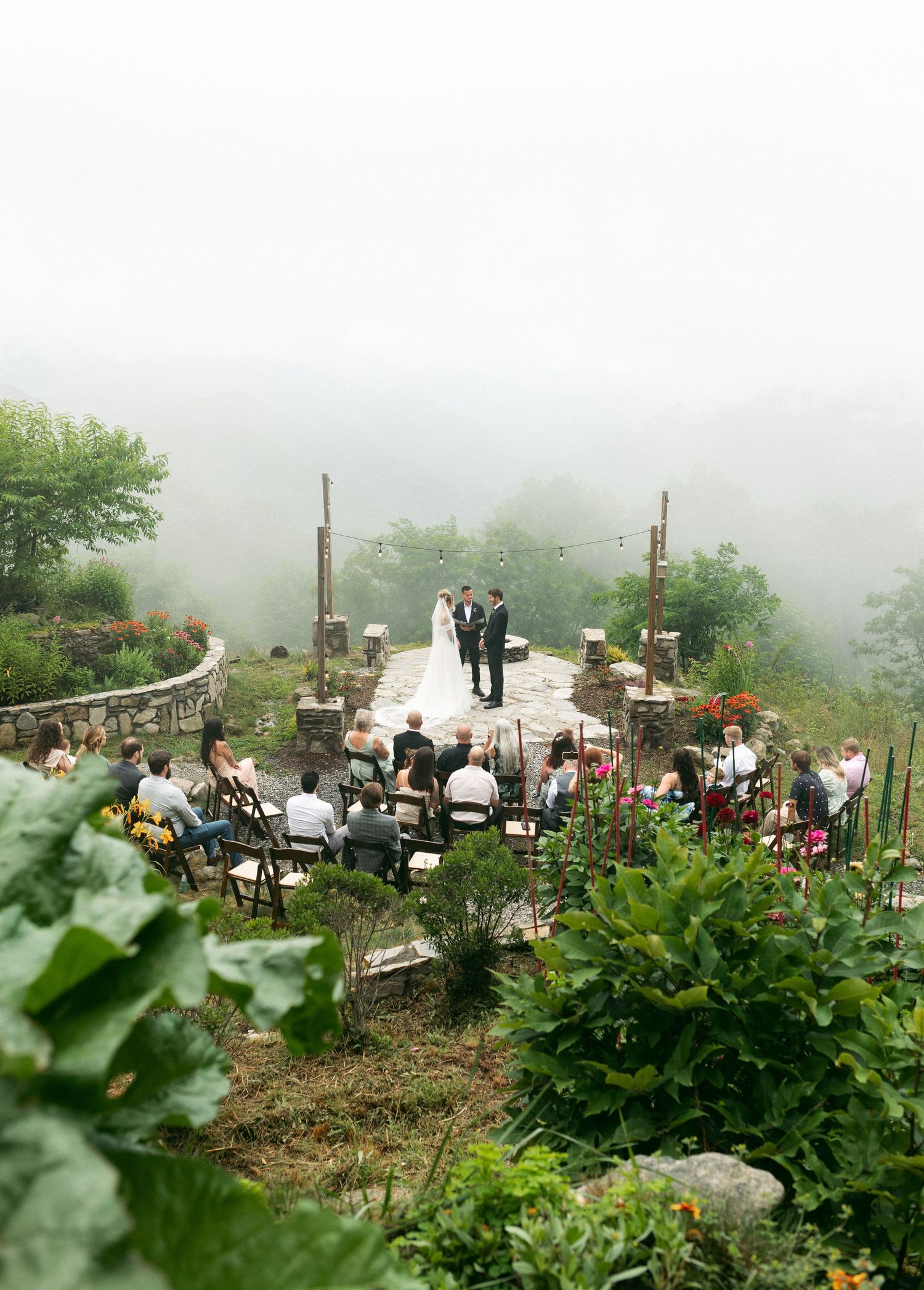 Misty mountain wedding ceremony on stone terrace with couple at altar surrounded by lush greenery and seated guests.