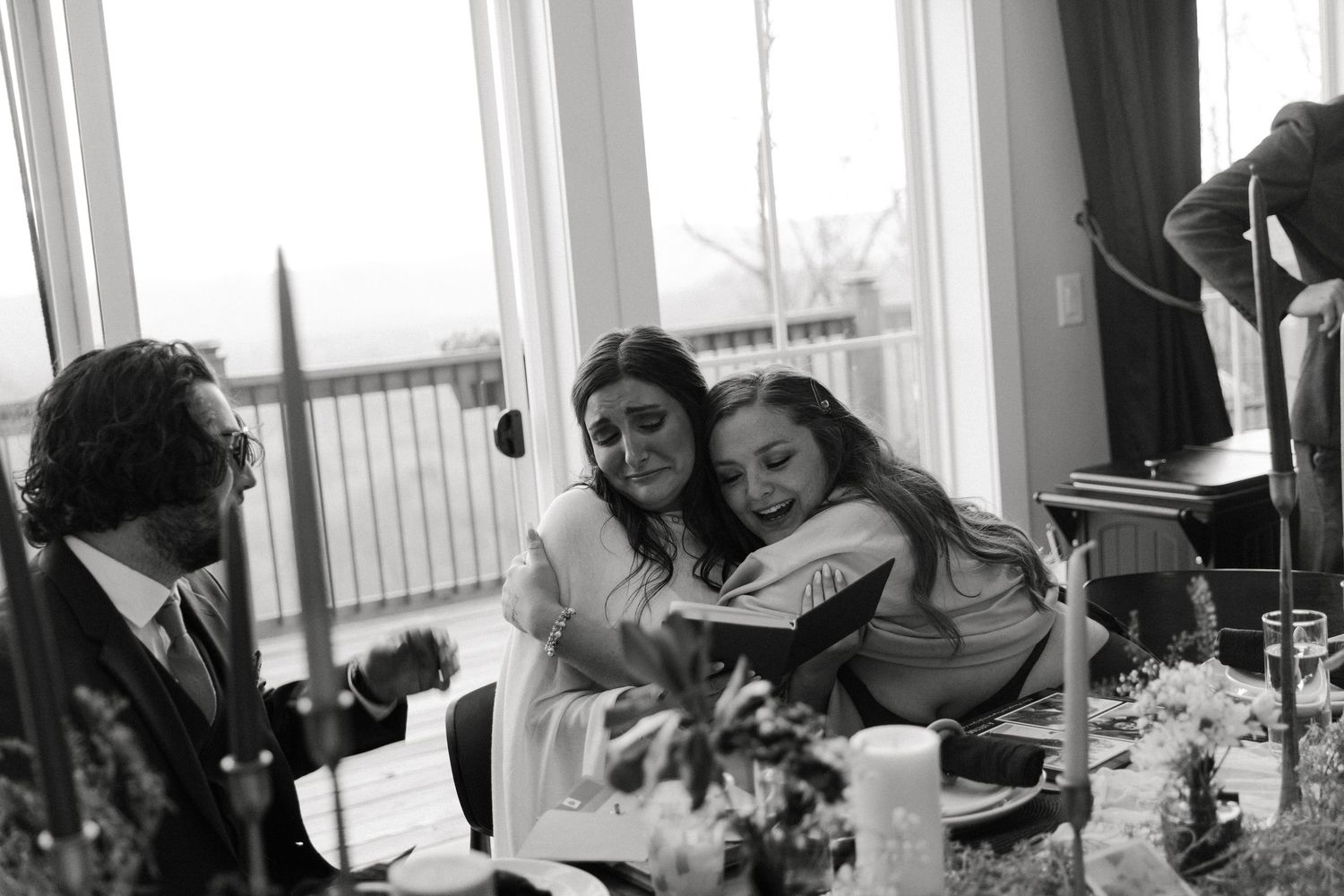 Two women share an emotional hug while reading at a wedding reception table in black and white photo.