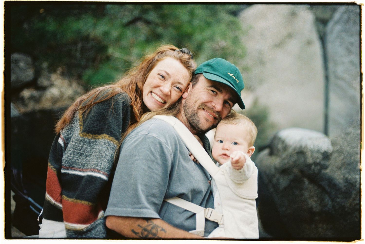 Happy family with smiling parents and baby in carrier posing outdoors near rocks in casual clothing.