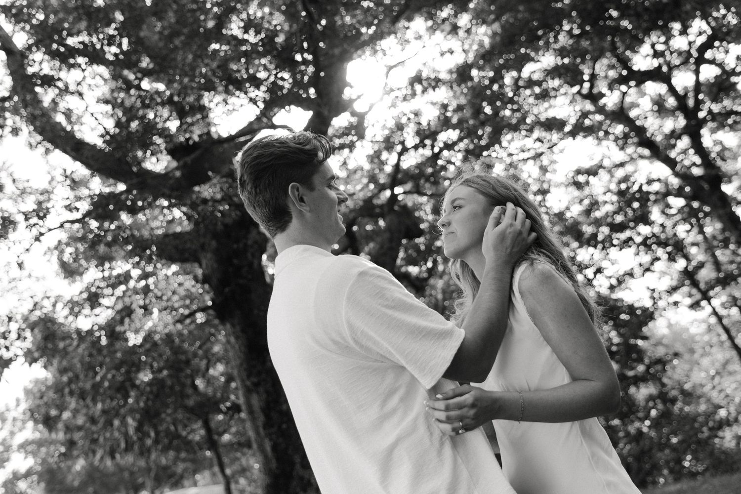Black and white photo of a romantic couple embracing outdoors under trees in soft natural light.