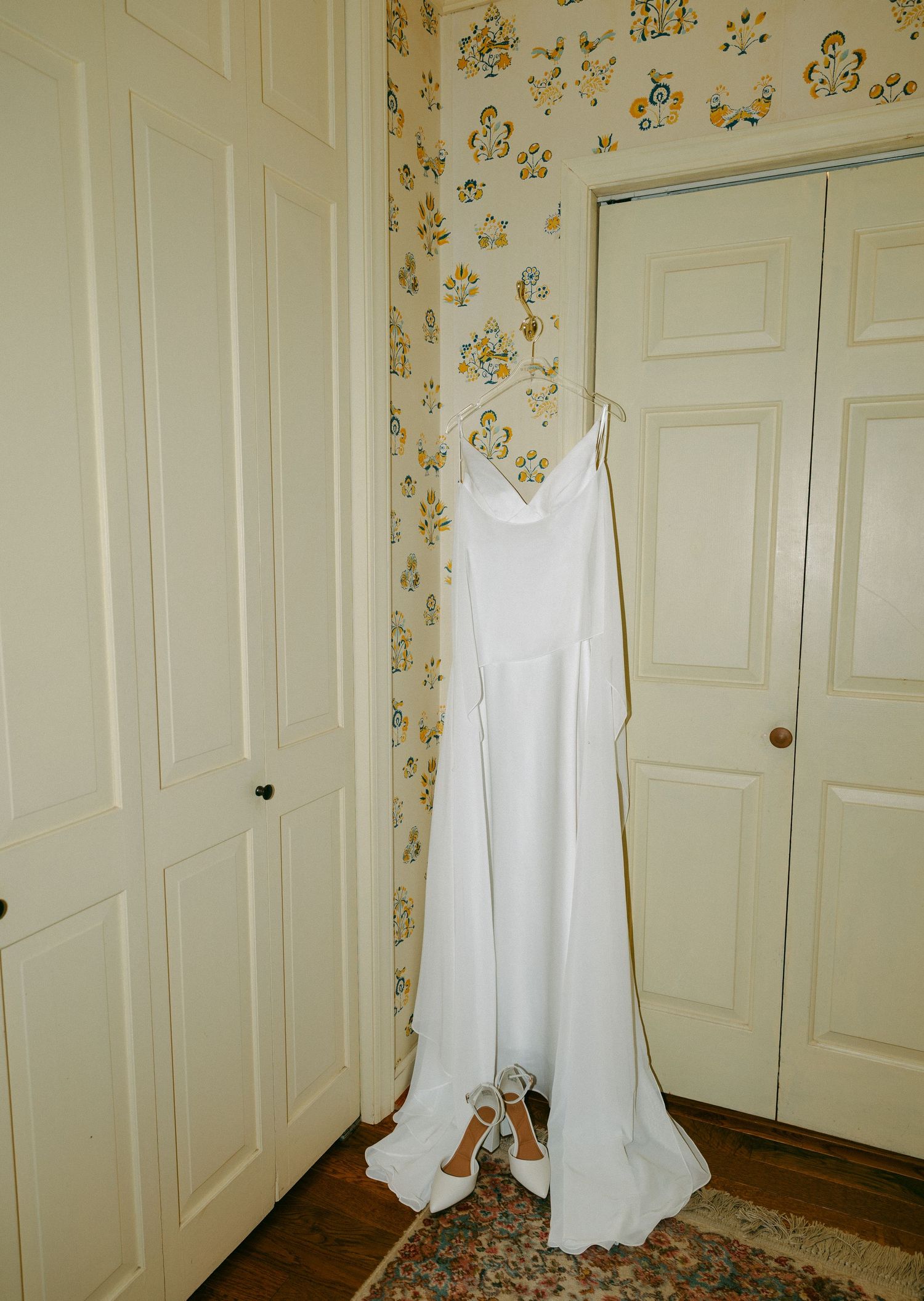 White wedding gown with train hanging on a door with floral wallpaper and heels below.