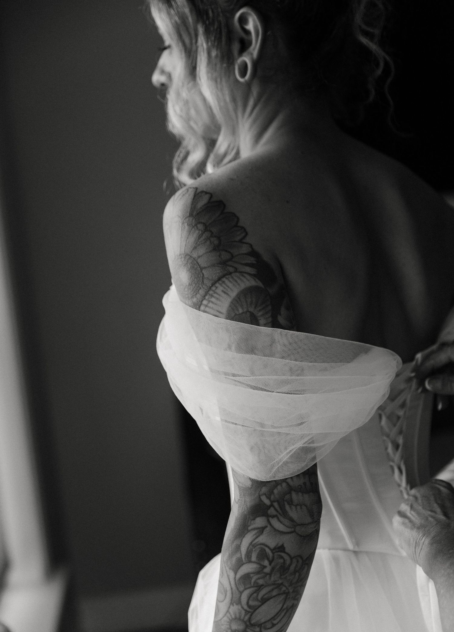 Black and white photo of a tattooed bride in an off-shoulder white wedding dress, showcasing intricate floral arm tattoos.