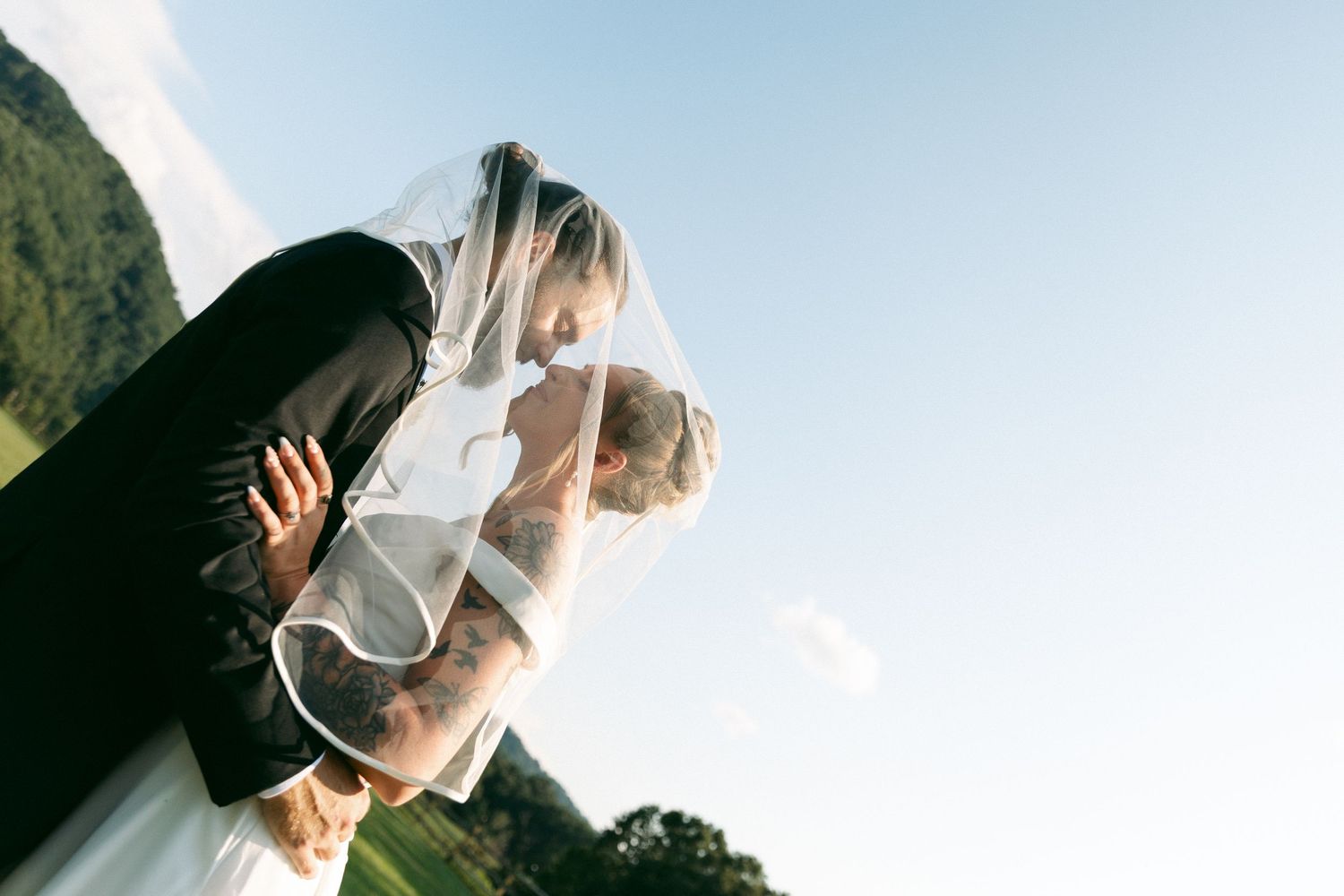 Bride and groom sharing a romantic kiss under a wedding veil outdoors with green hills and blue sky.