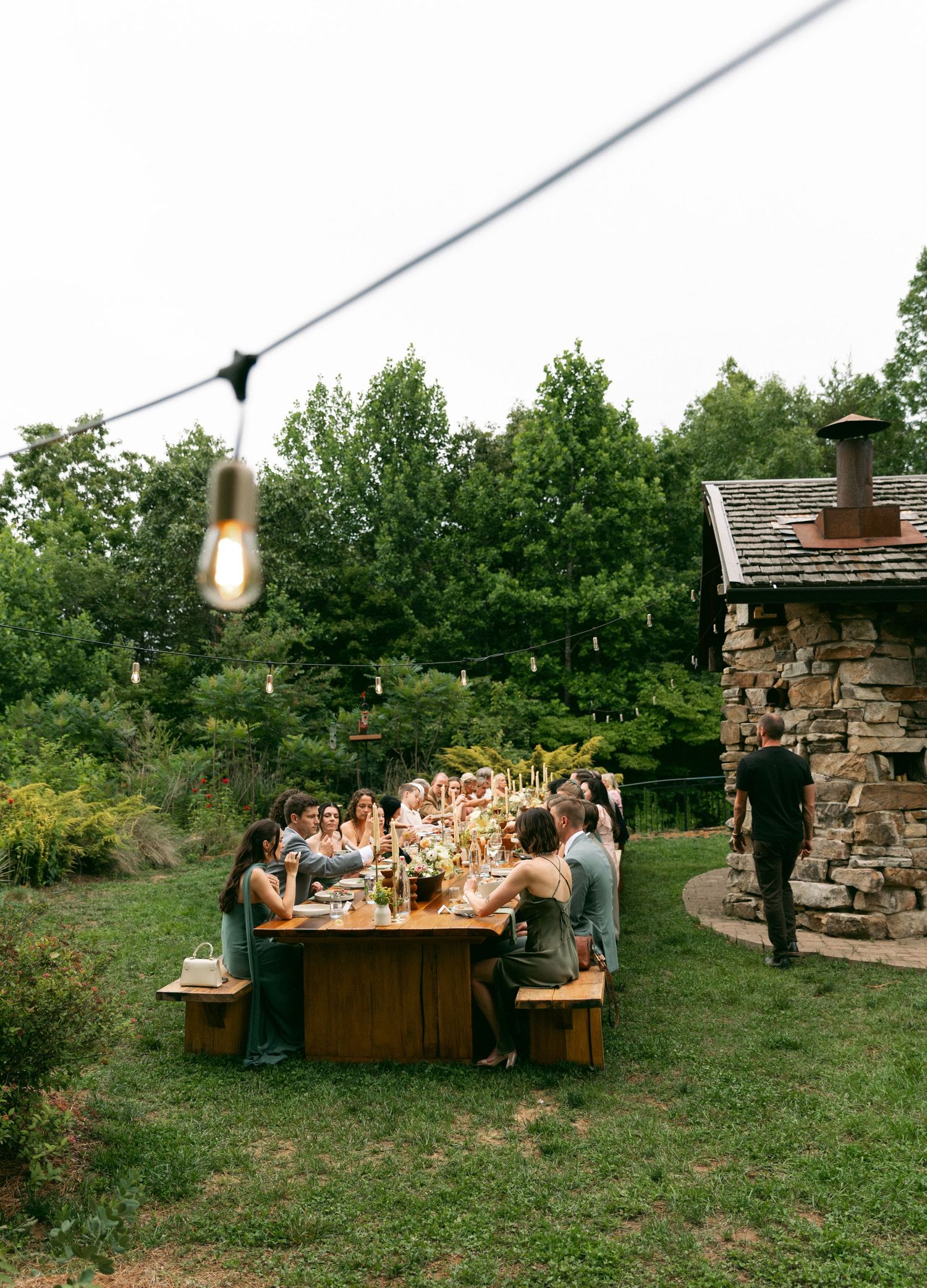 Group of people dining outdoors at a wooden table surrounded by lush greenery with string lights and a stone building nearby.