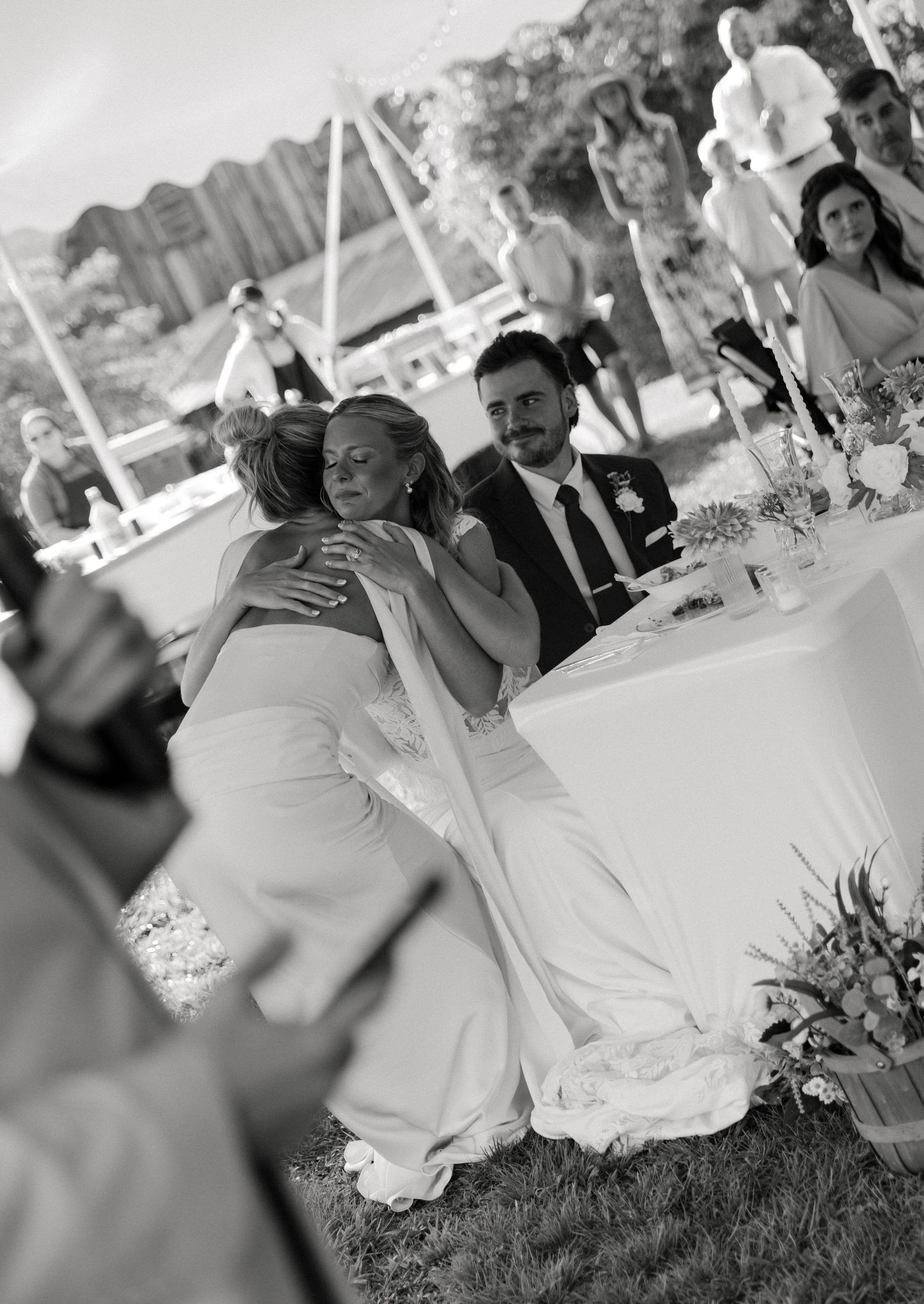 Black and white wedding photo of bride hugging guest while groom smiles at outdoor reception table.