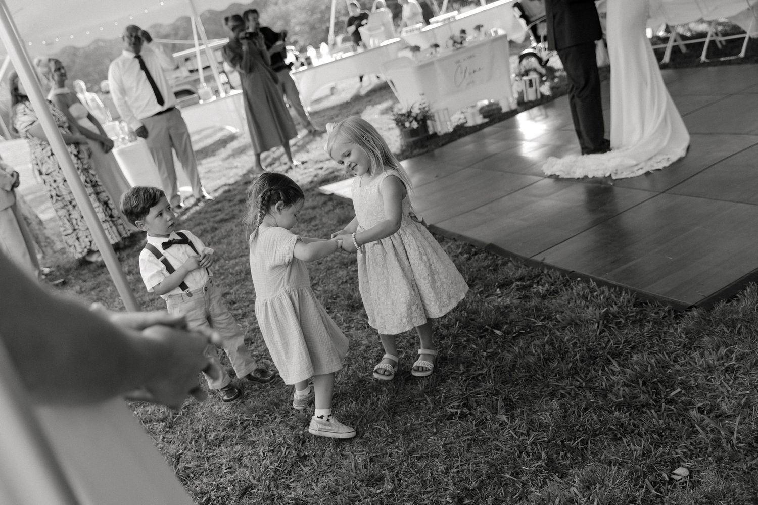 Two flower girls in white dresses dance together on grass at an outdoor wedding reception under a tent.