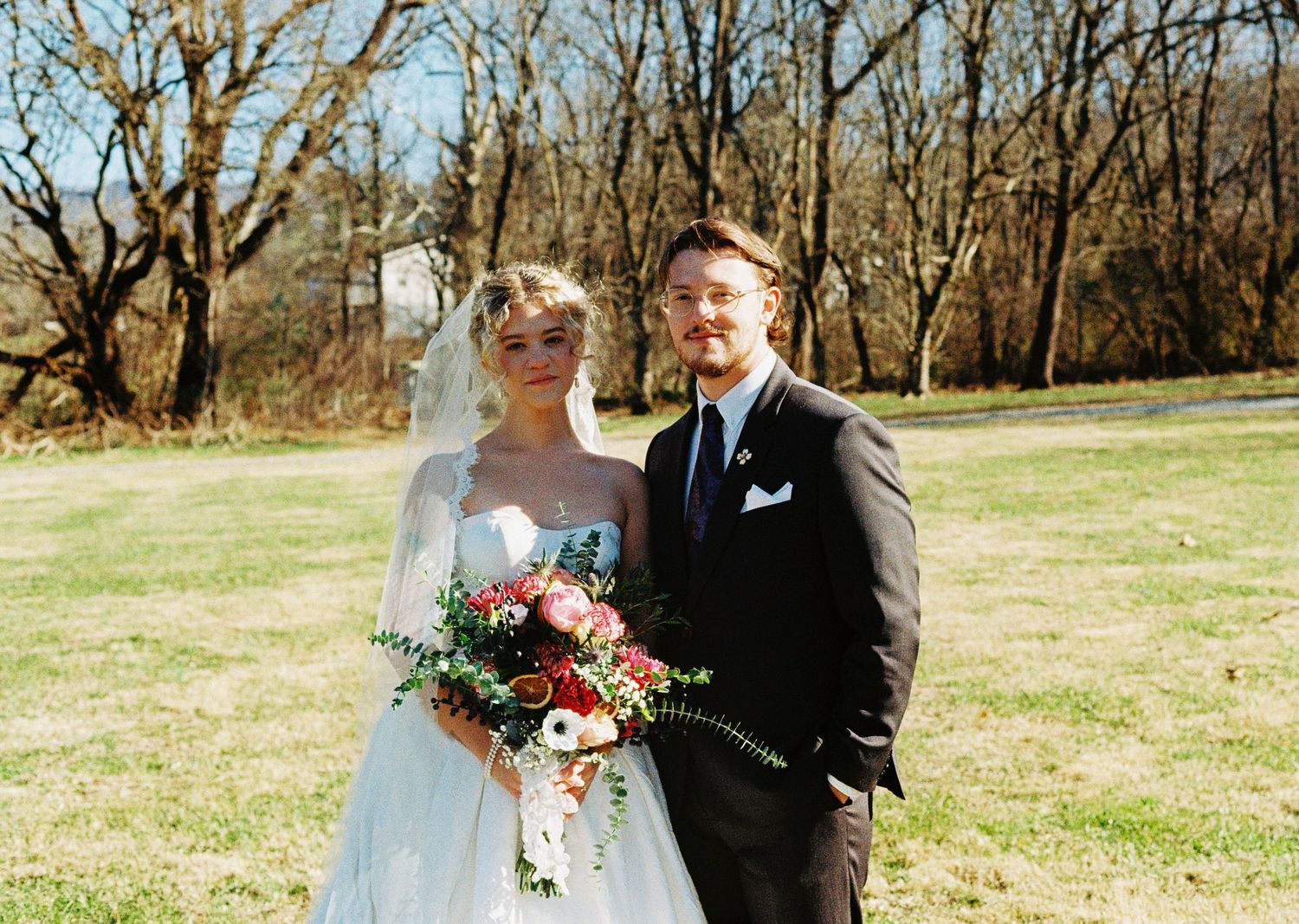 Bride and groom posing outdoors on green grass, bride in white gown with veil holding colorful bouquet.