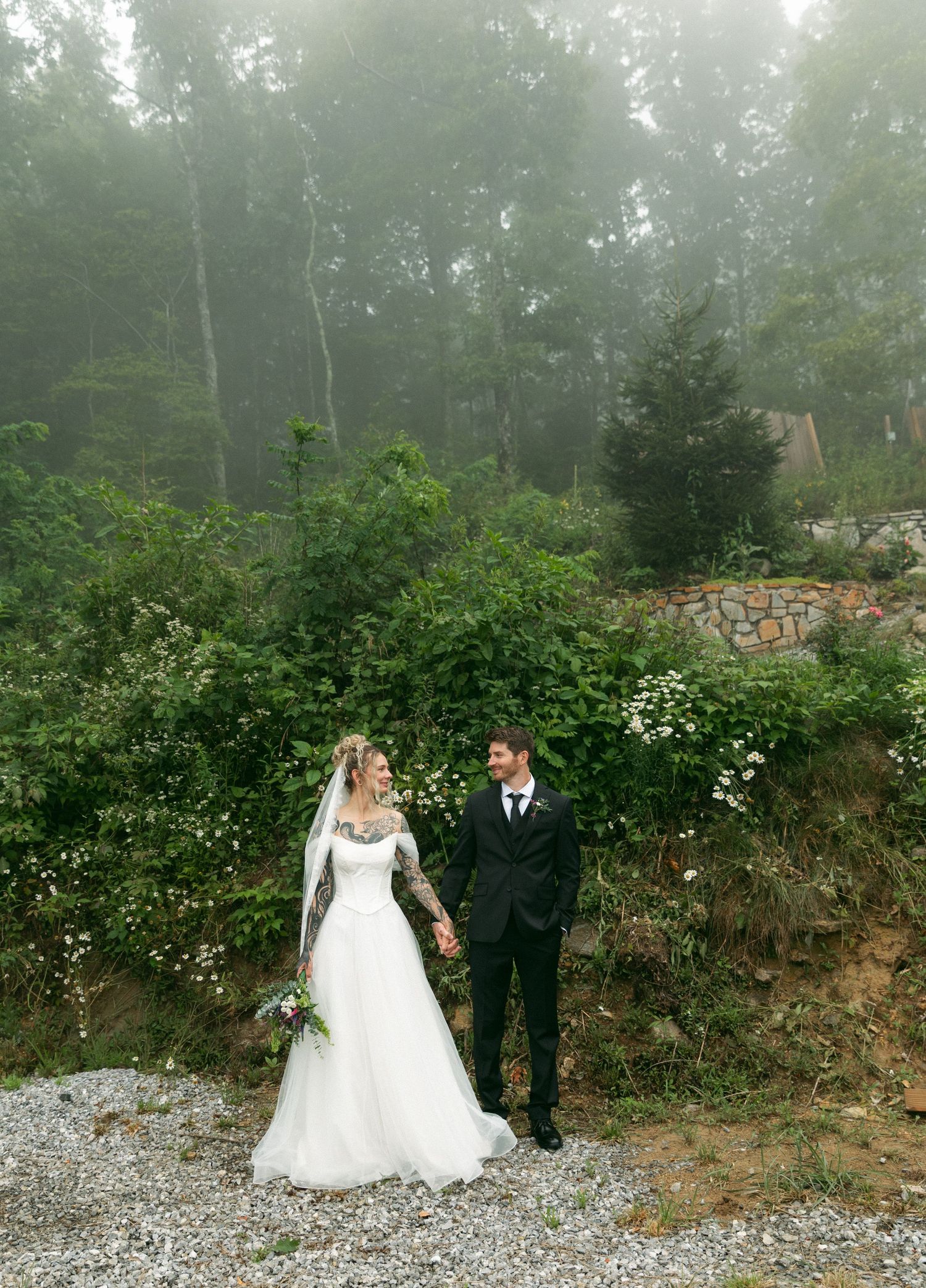 Bride and groom hold hands in a misty forest setting, bride wearing lace long-sleeve gown holding bouquet.