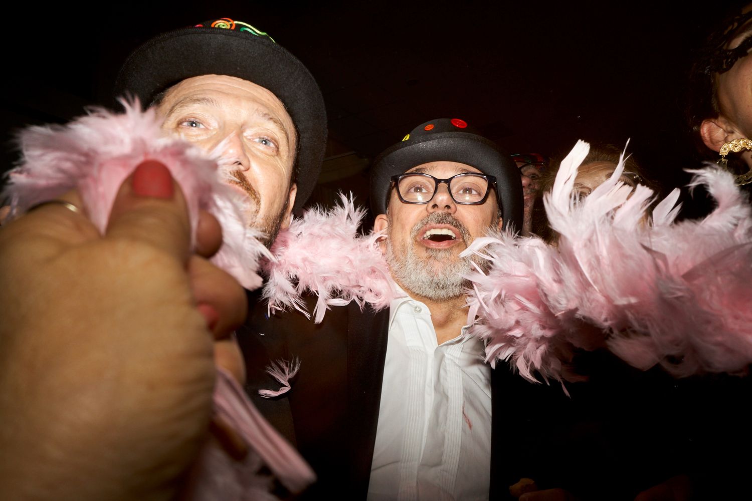 Invitados disfrazados con boas de plumas rosas y sombreros de fiesta riendo en primer plano