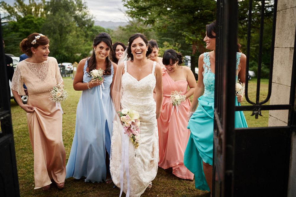 Novia sonriente caminando junto a sus damas de honor con vestidos en tonos pastel, cruzando una reja hacia el exterior