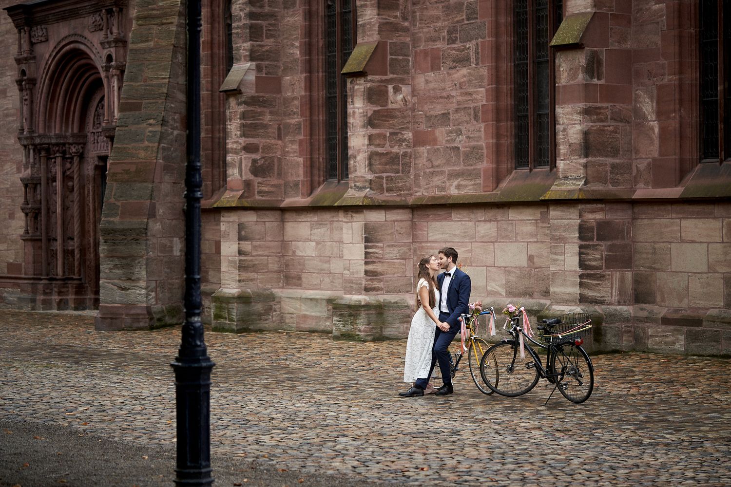 Novios a punto de besarse junto a sus bicicletas decoradas con flores frente a una iglesia de piedra