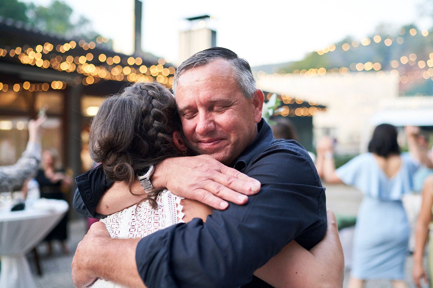 Padre de la novia abrazando emocionadola durante la fiesta al aire libre, con luces colgantes de fondo