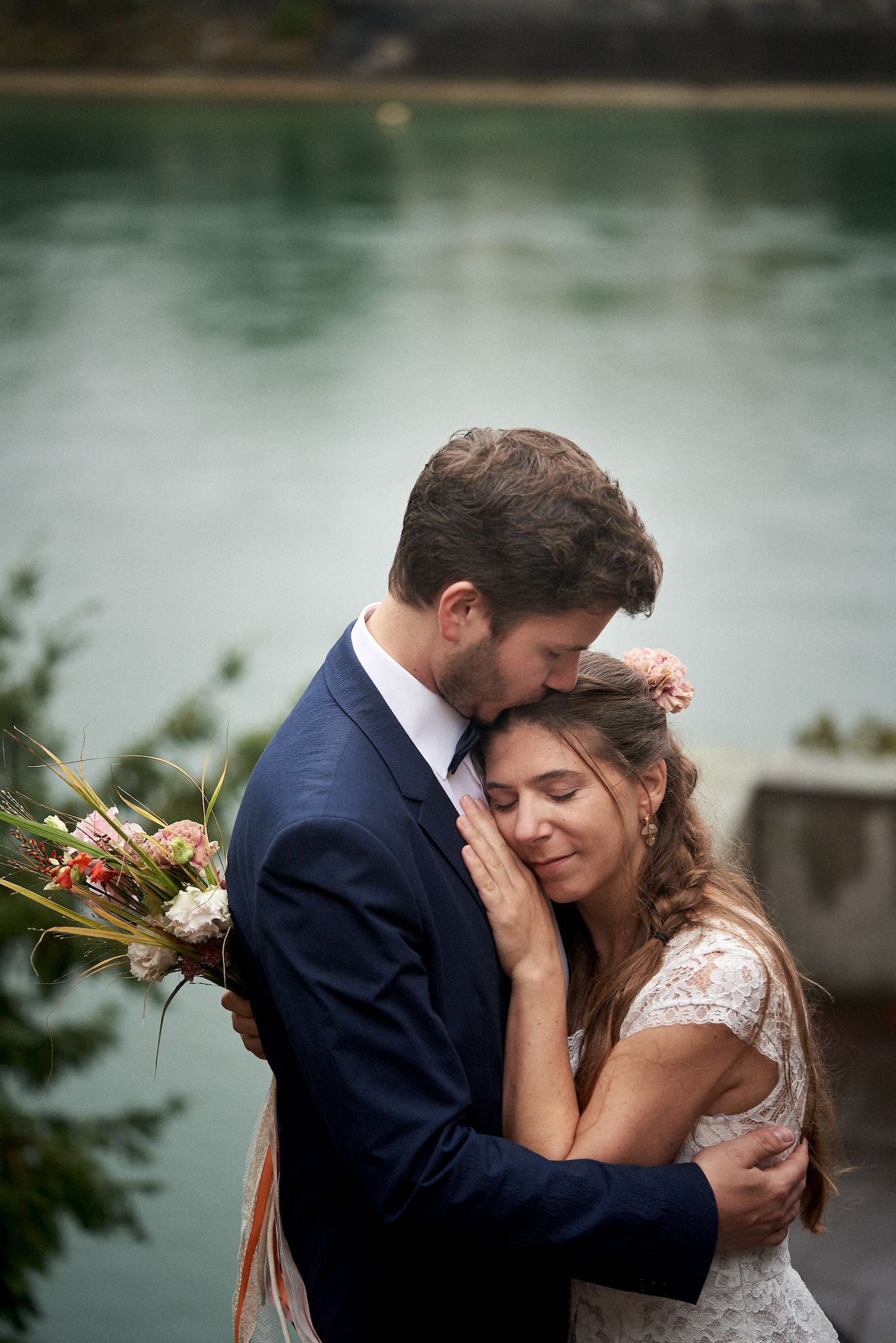 Novios abrazados junto a un lago, el novio besando la frente de la novia que sostiene un ramo de flores