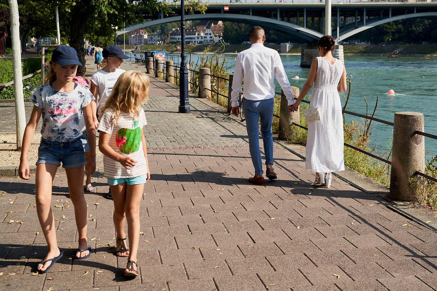 Pareja de novios caminando junto a un río tomados de la mano, con un puente de fondo, mientras niños curiosos los miran