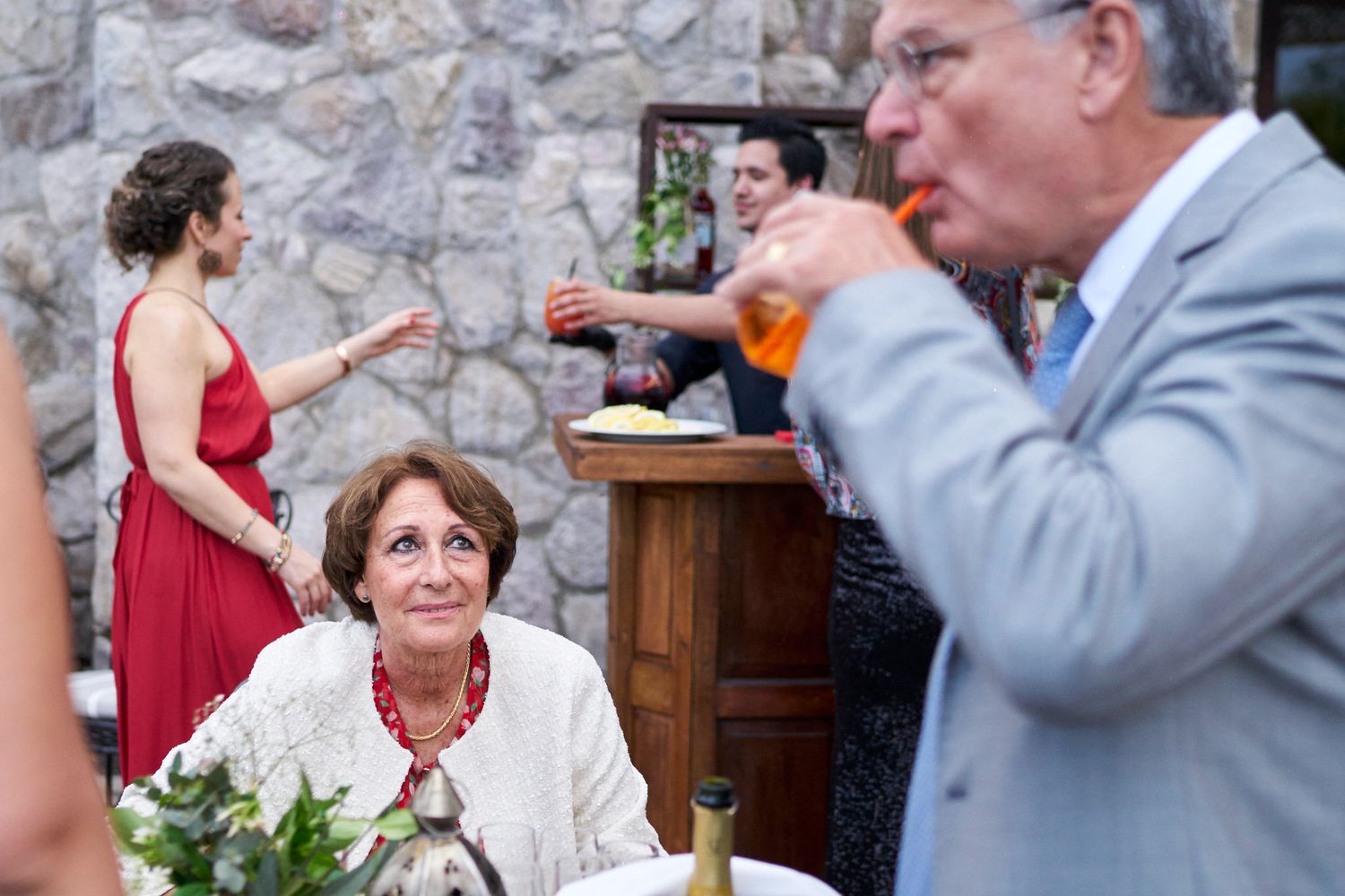 Invitado mayor bebiendo con una pajita mientras otros invitados conversan de fondo. Una mujer lo observa.