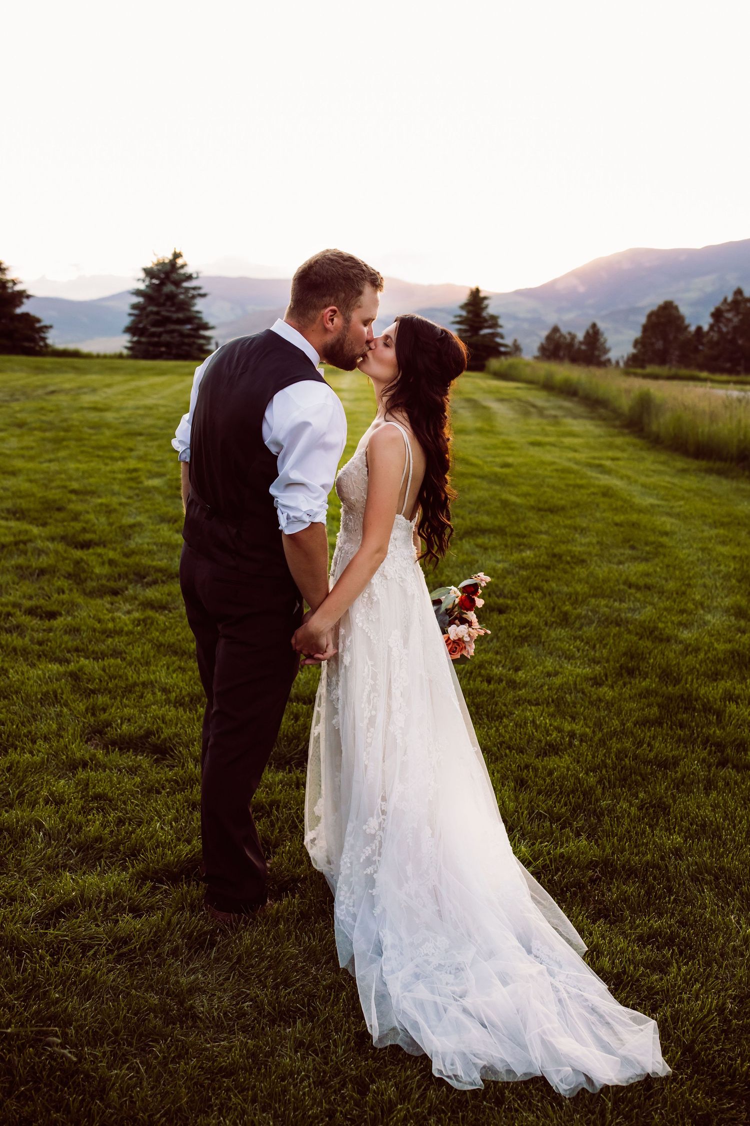 Bride and groom share a romantic kiss at sunset on a lush green field with mountain views on their wedding day.