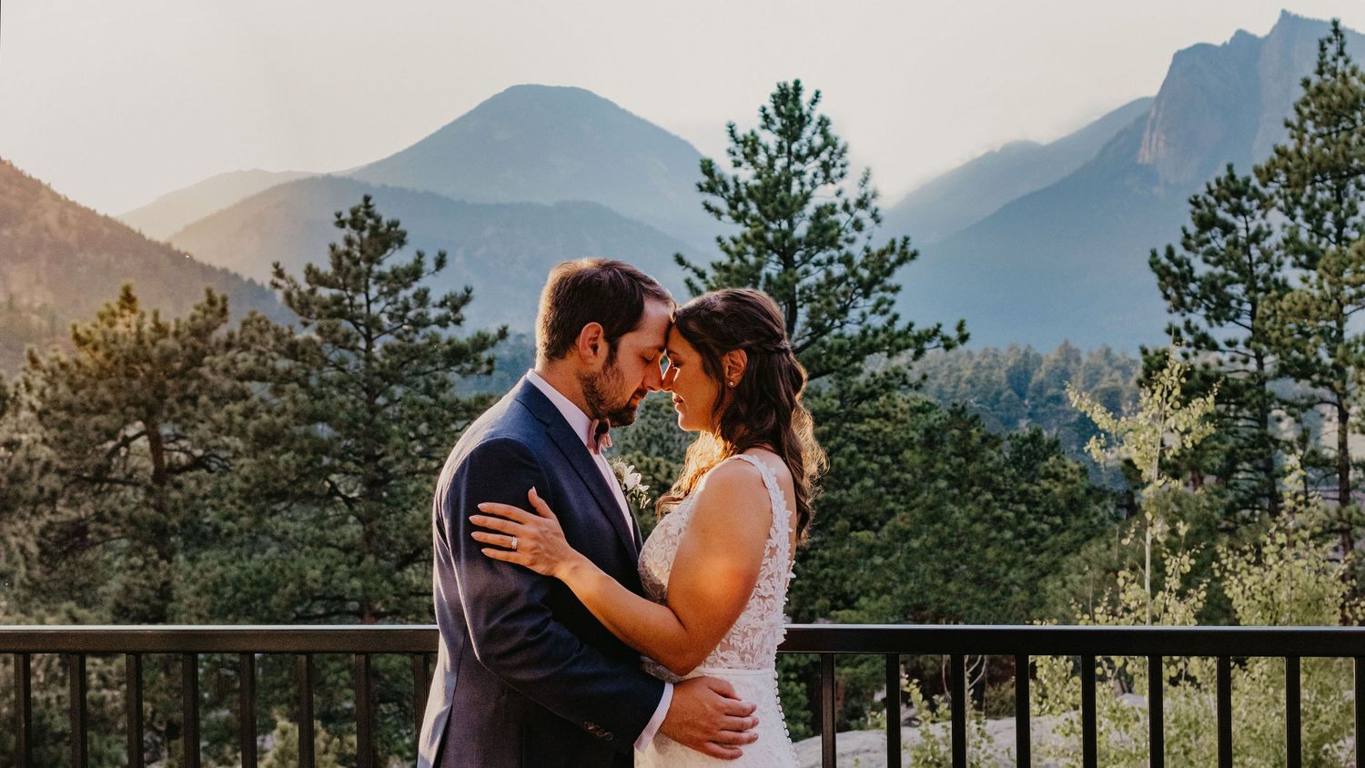 A bride and groom share a romantic moment on a mountain balcony with scenic pine trees and peaks at sunset.